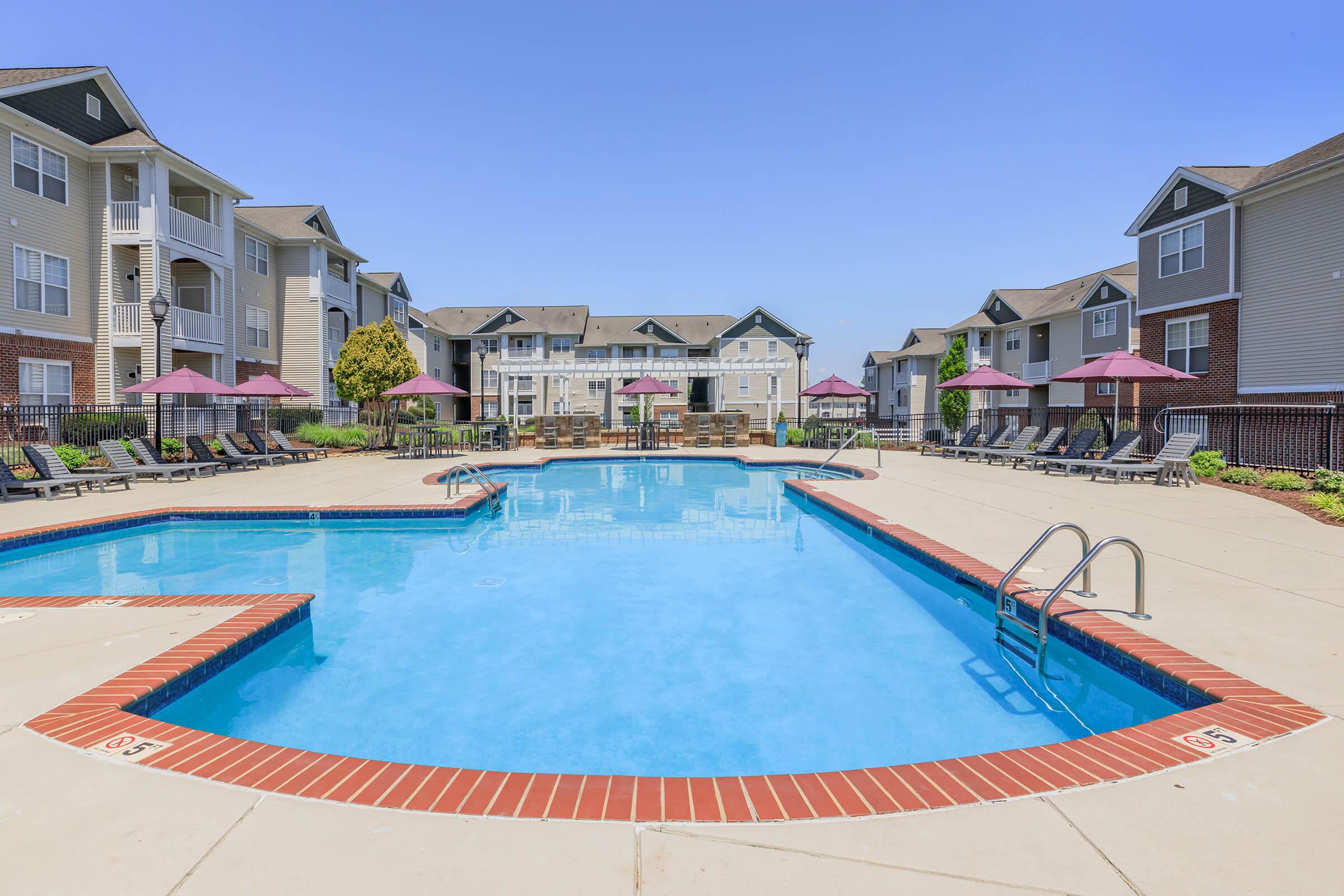 A clear blue swimming pool surrounded by lounge chairs and umbrellas, with modern apartment buildings in the background. The sky is bright and clear, creating a relaxing atmosphere for leisure and enjoyment.