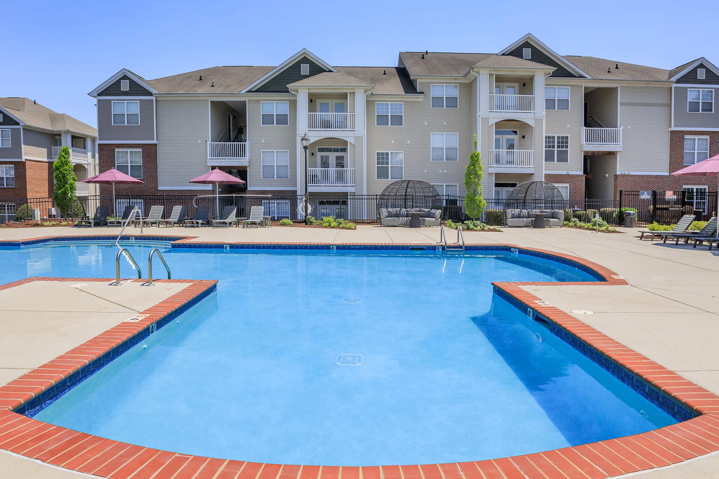 A sunny outdoor swimming pool surrounded by lounge chairs and shaded by pink umbrellas. In the background, there are multi-story residential buildings with balconies. The pool features a brick border and clear blue water, creating a relaxing atmosphere for residents.