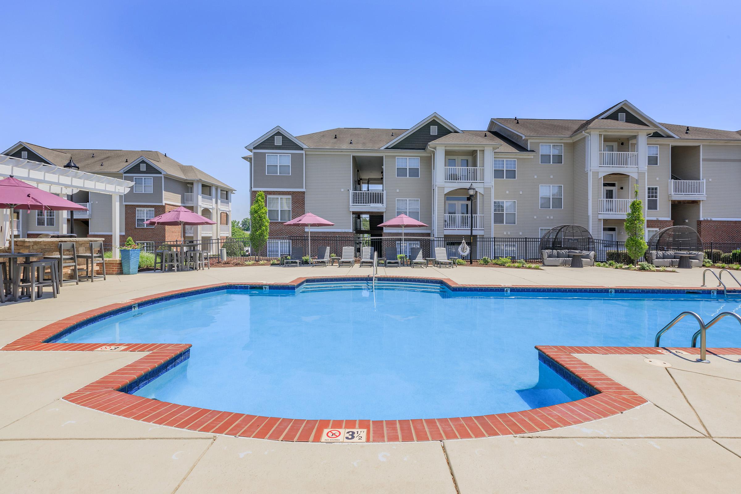 A residential apartment complex featuring a clear blue swimming pool surrounded by lounge chairs and umbrellas. The pool area is bordered by well-maintained landscaping and buildings with balconies. The sky is bright and clear, indicating a sunny day.