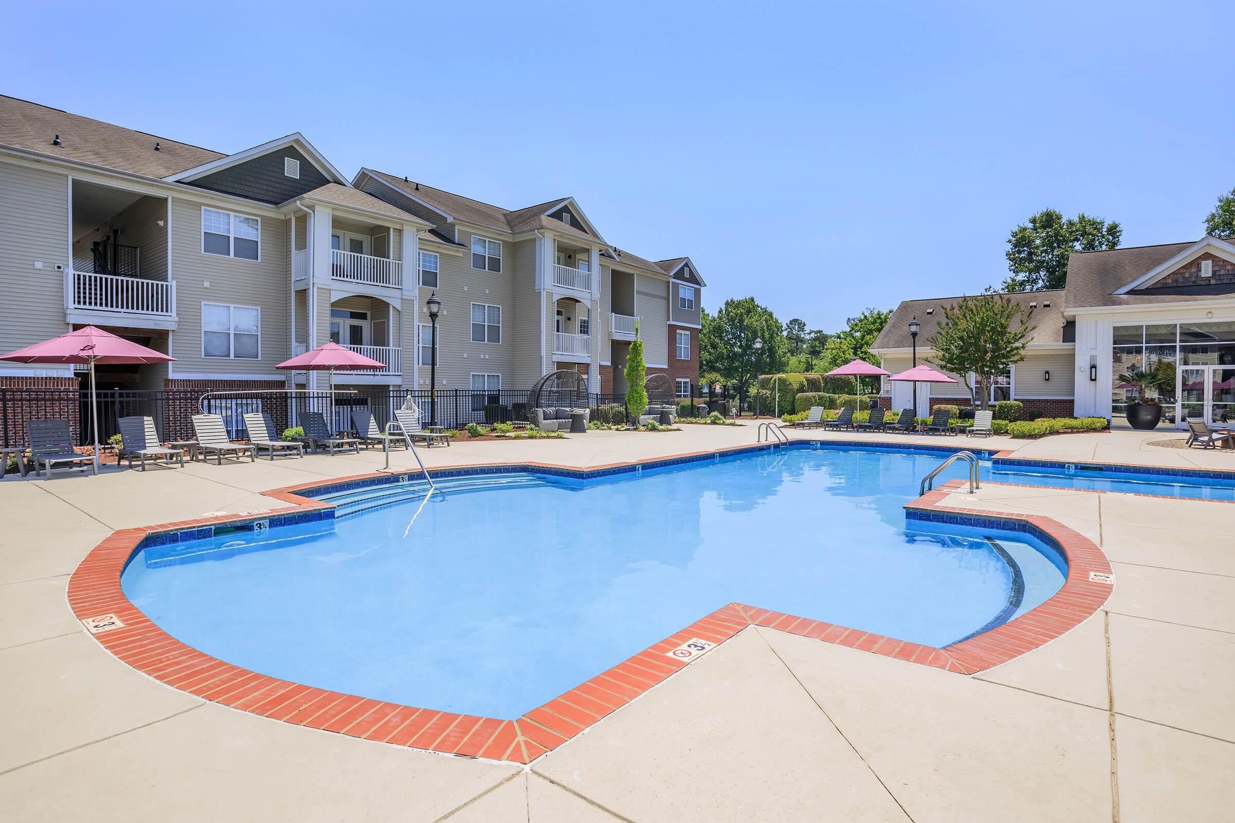 A clear blue swimming pool in a sunny residential area, surrounded by lounge chairs and shaded by pink umbrellas. The pool is bordered by a decorative red brick edge, with modern apartment buildings visible in the background. Well-maintained landscaping enhances the inviting atmosphere.