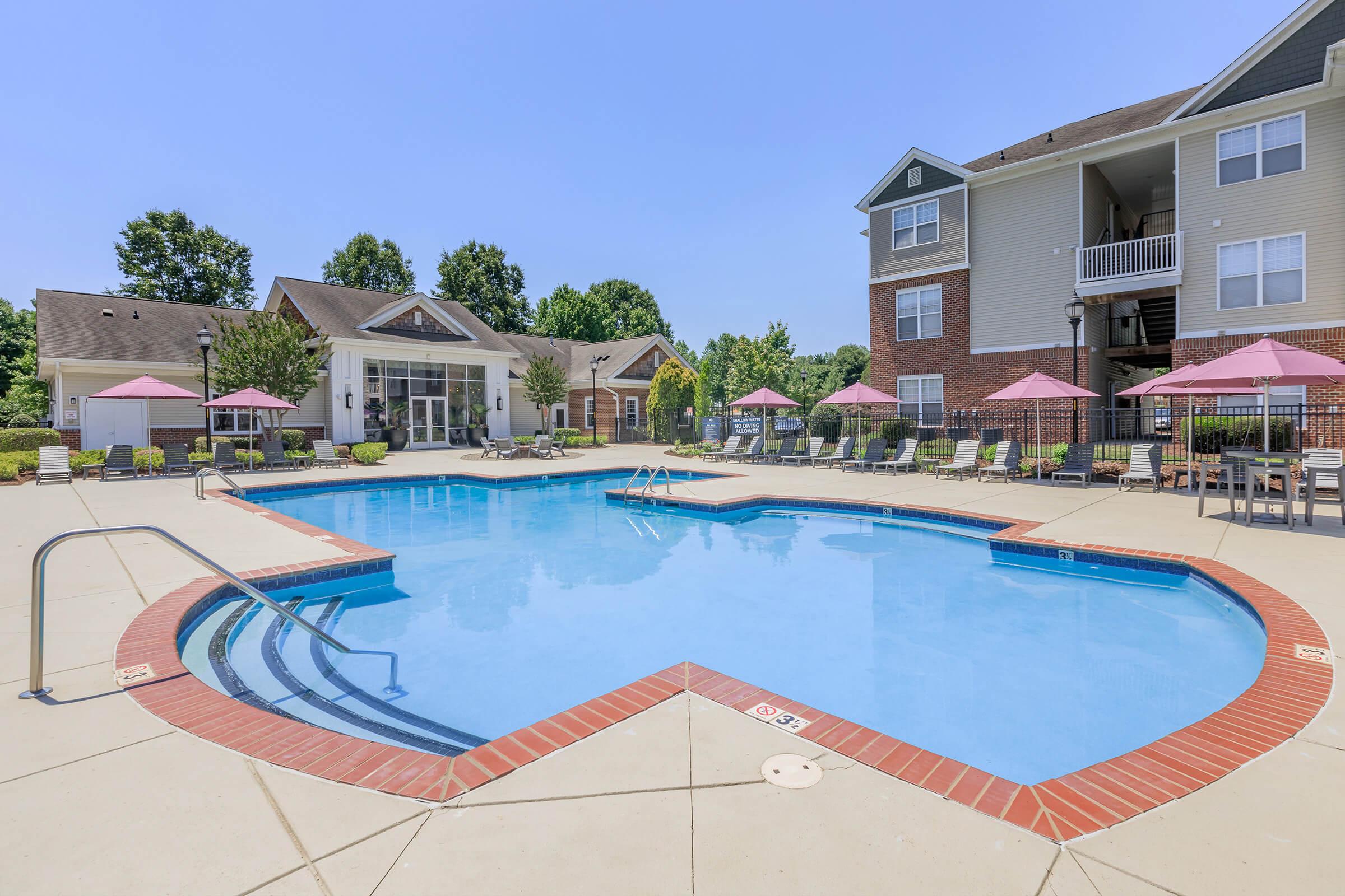A clear blue swimming pool surrounded by lounge chairs and pink umbrellas, with a landscaped area and a multi-story residential building in the background under a bright blue sky. The scene evokes a relaxing outdoor atmosphere perfect for leisure and recreation.