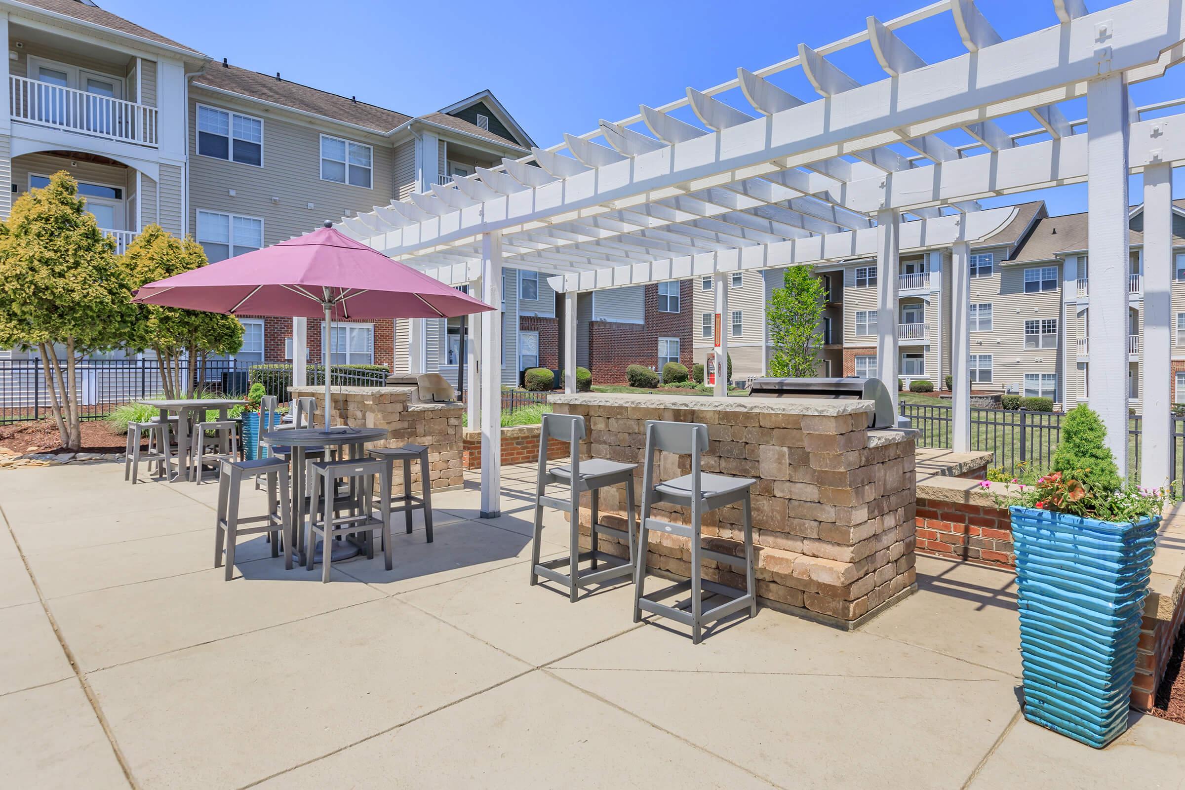 Outdoor lounge area featuring a stone bar with high stools, a pink umbrella, and tables. Surrounded by green landscaping and modern apartment buildings under a clear blue sky. Ideal for relaxation and social gatherings.
