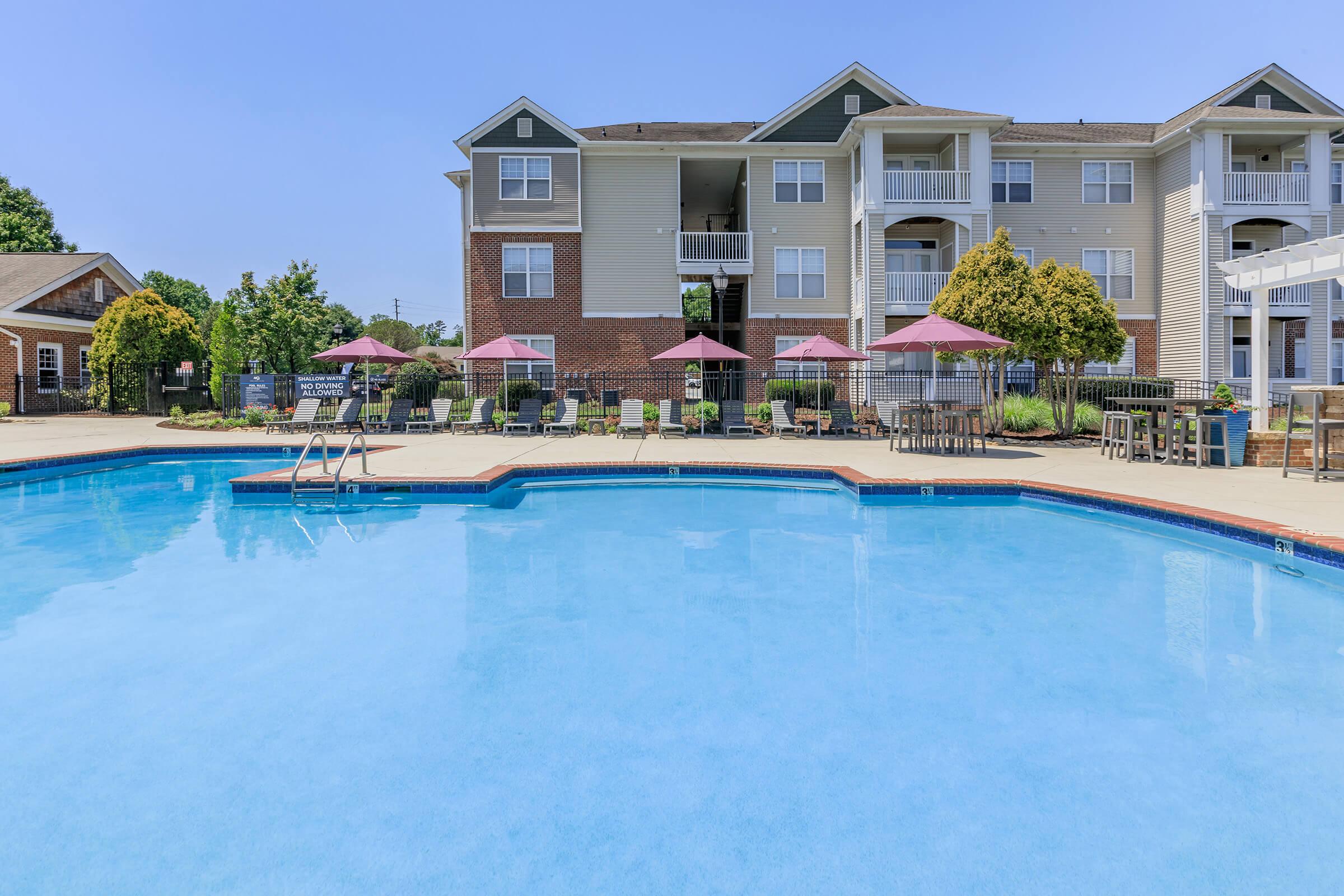 A sunny outdoor pool area featuring clear blue water, surrounded by lounge chairs and umbrellas. The background includes a modern apartment building with balconies, lush greenery, and a well-maintained landscape, creating an inviting atmosphere for relaxation and leisure.