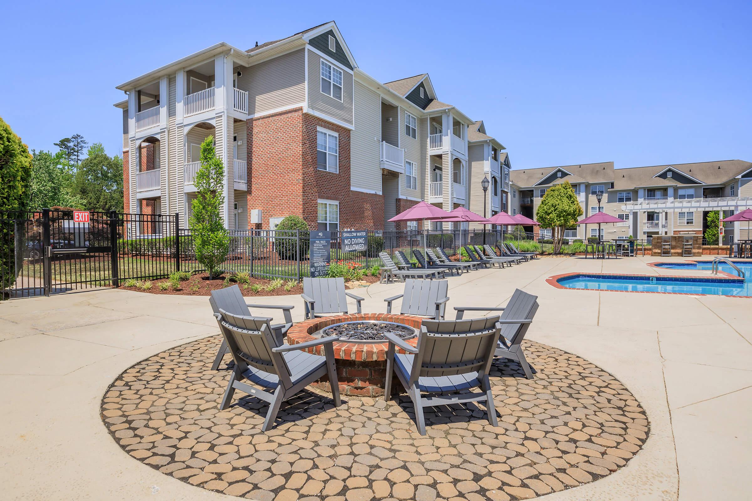A sunny outdoor area featuring a circular stone patio with several gray chairs arranged around a fire pit. In the background, a residential apartment complex with multiple stories and balconies can be seen. There is a swimming pool nearby, surrounded by pink umbrellas and lounge chairs.