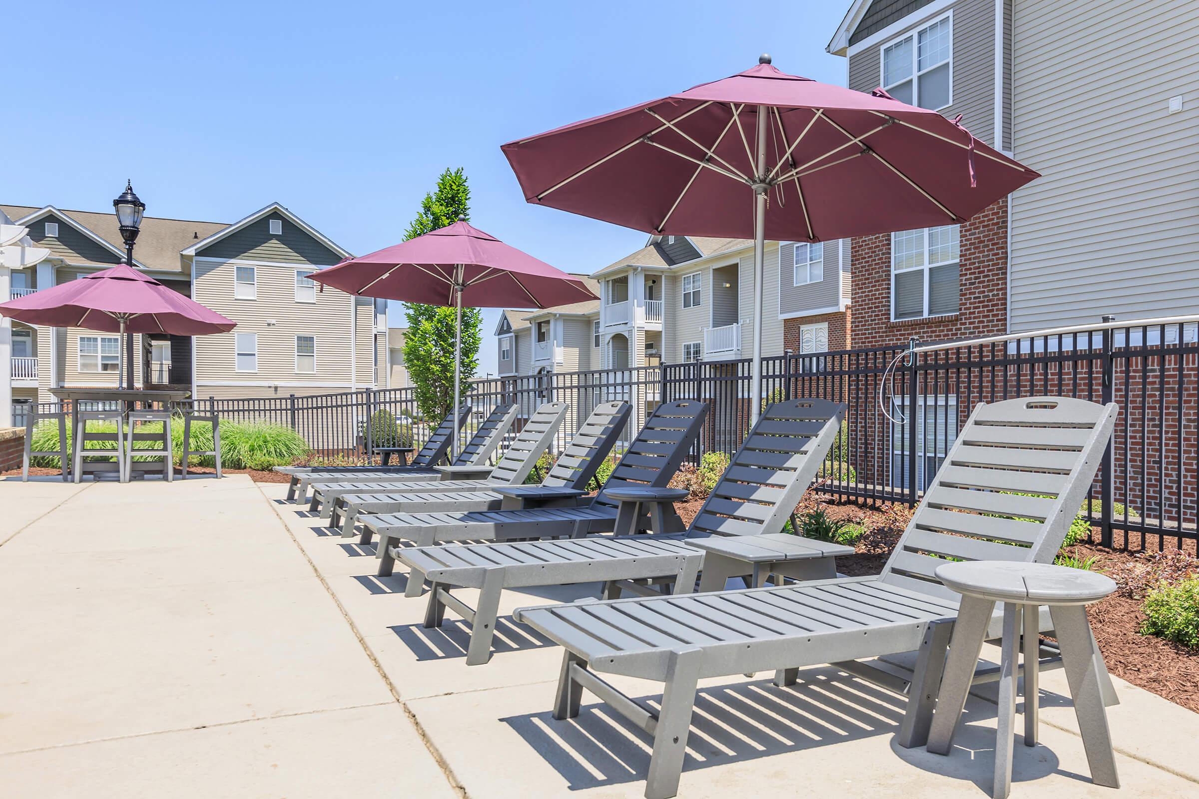 Loungers with maroon umbrellas are positioned around a poolside area. The scene features a landscaped courtyard with apartment buildings in the background under a clear blue sky.