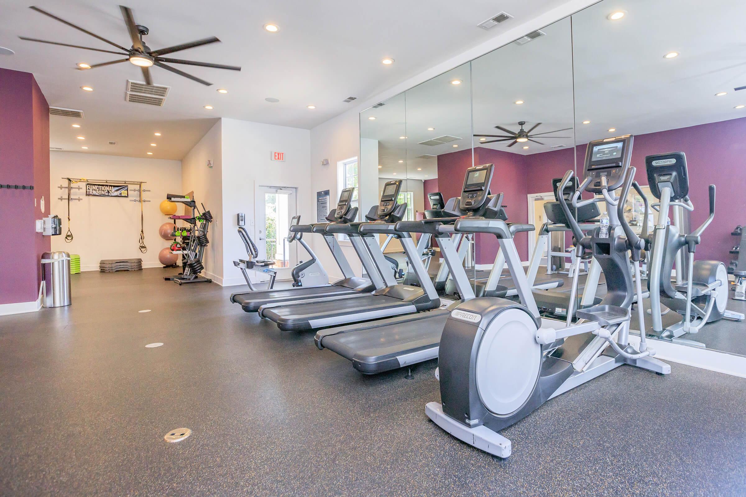 A bright and spacious gym with several treadmills and elliptical machines facing a large mirror. The walls are painted in purple, and there are various exercise equipment and weights on the side. A ceiling fan is visible, contributing to a comfortable workout environment.