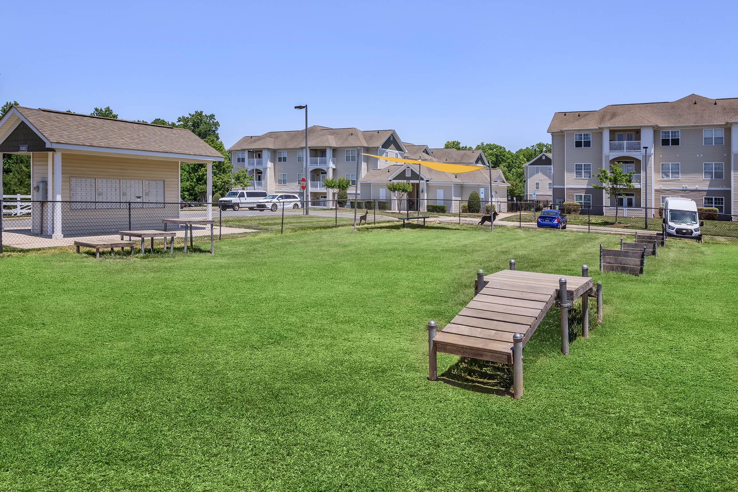 A spacious green lawn with a wooden pathway leading to a fenced area. In the background, there are two multi-story apartment buildings with balconies and well-maintained landscaping. A parking area is visible with parked cars. Clear blue sky above.