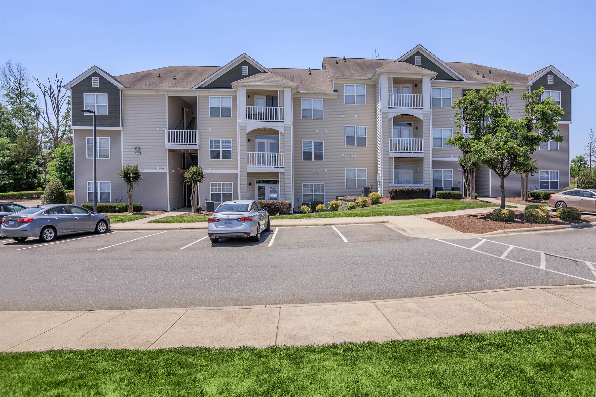 Modern apartment building with three stories, featuring a light gray exterior and multiple balconies. The surrounding area includes landscaped lawns and a parking lot with several cars. Clear blue sky above the building, creating a bright and welcoming atmosphere.