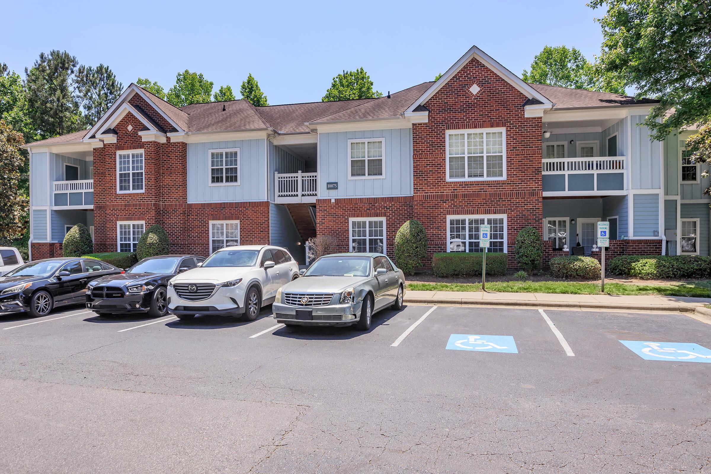 A well-maintained apartment complex with two levels, featuring brick and blue siding. The front has several parking spaces, including handicapped spots. Lush green landscaping surrounds the building, providing a pleasant atmosphere on a sunny day.