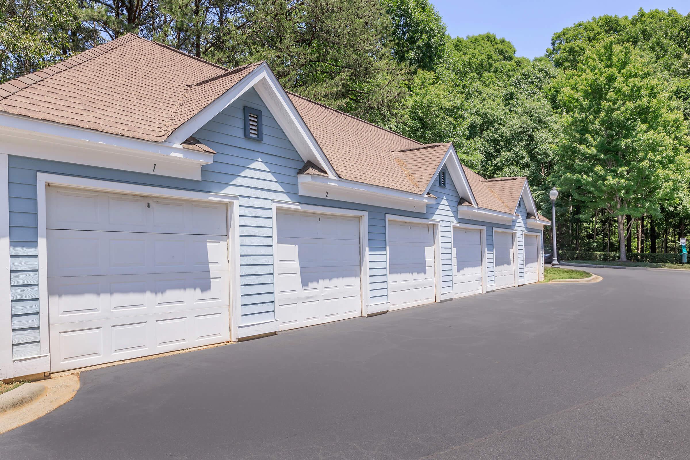 Row of blue garages with white doors, lined up along a newly paved road. Green trees in the background provide a natural setting. The scene is bright and well-maintained, showcasing a peaceful, suburban environment.