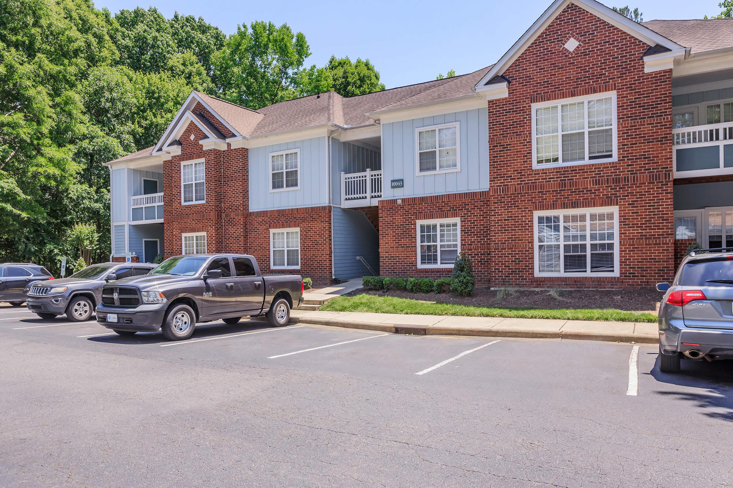 A modern multi-unit apartment building with a mix of brick and blue exterior. The building features balconies and several windows. In the foreground, parked cars occupy a paved lot, surrounded by greenery and trees. Clear blue skies are visible, indicating a sunny day.
