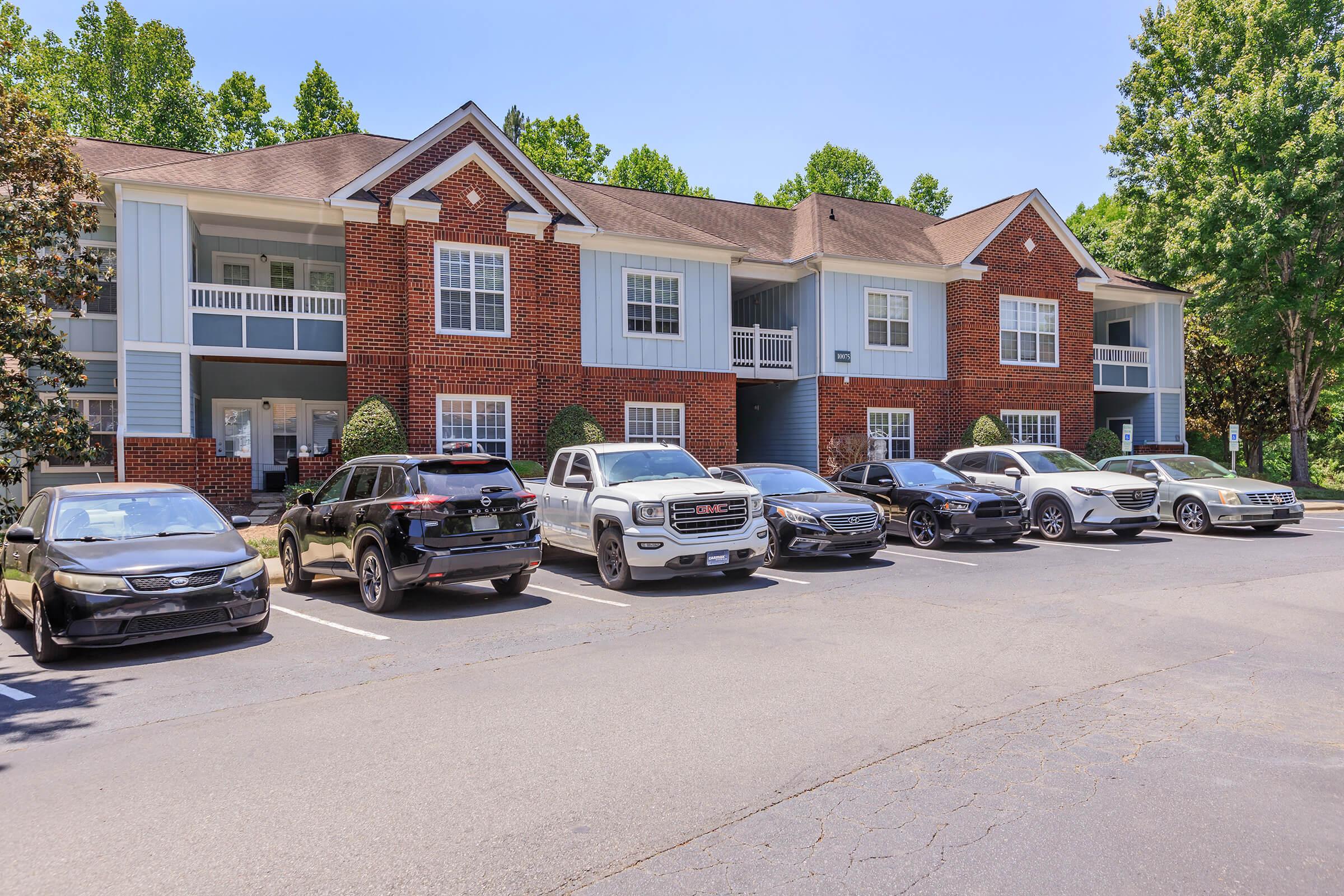 Row of parked vehicles in front of a multi-story apartment building. The building features a combination of brick and blue siding, with balconies visible on the upper level. Lush green trees provide landscaping in the background, under a clear blue sky.