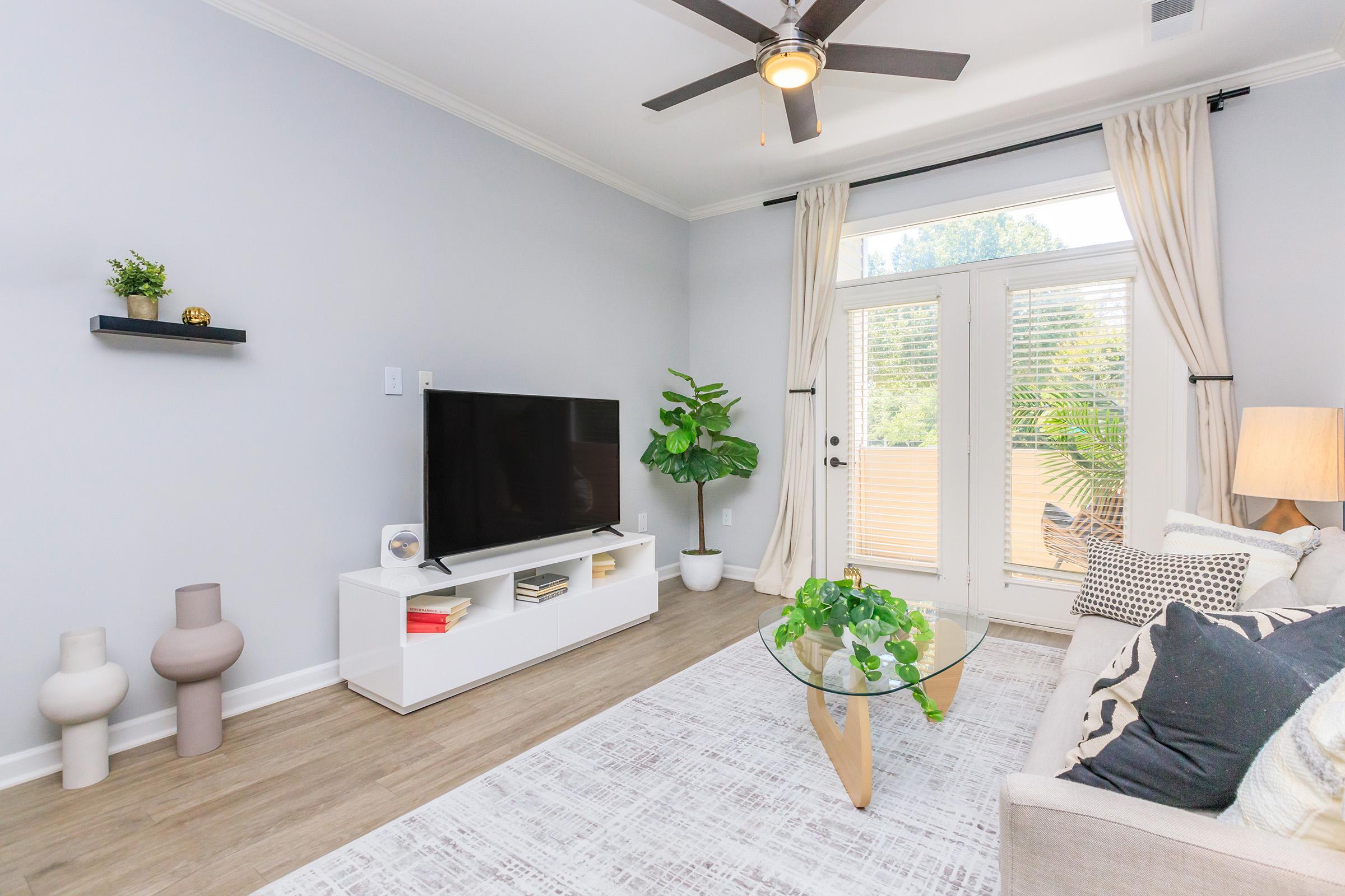 Bright and modern living room featuring a light-colored sofa, a glass coffee table with greenery, a flat-screen TV on a white media console, and large windows with sheer curtains. Decorative plants and minimalist decor enhance the inviting atmosphere. Wood flooring complements the neutral color scheme.