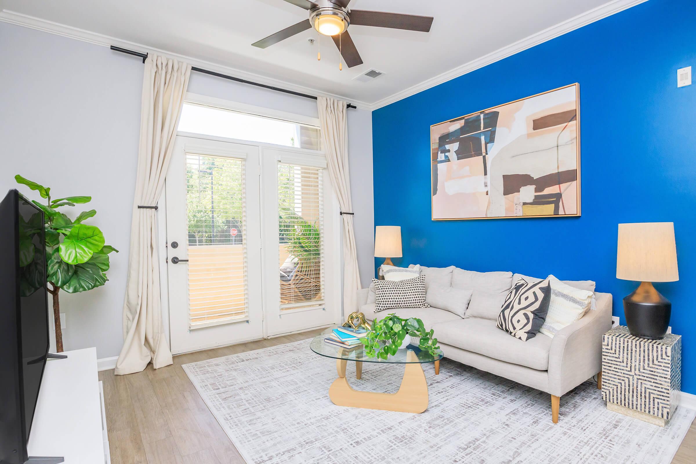 Modern living room with a light gray sofa adorned with patterned cushions, a round glass coffee table, and a decorative green plant. The walls feature a deep blue accent paint, while a large artwork hangs above the couch. Two stylish lamps illuminate the space, and double doors lead to an outdoor area.