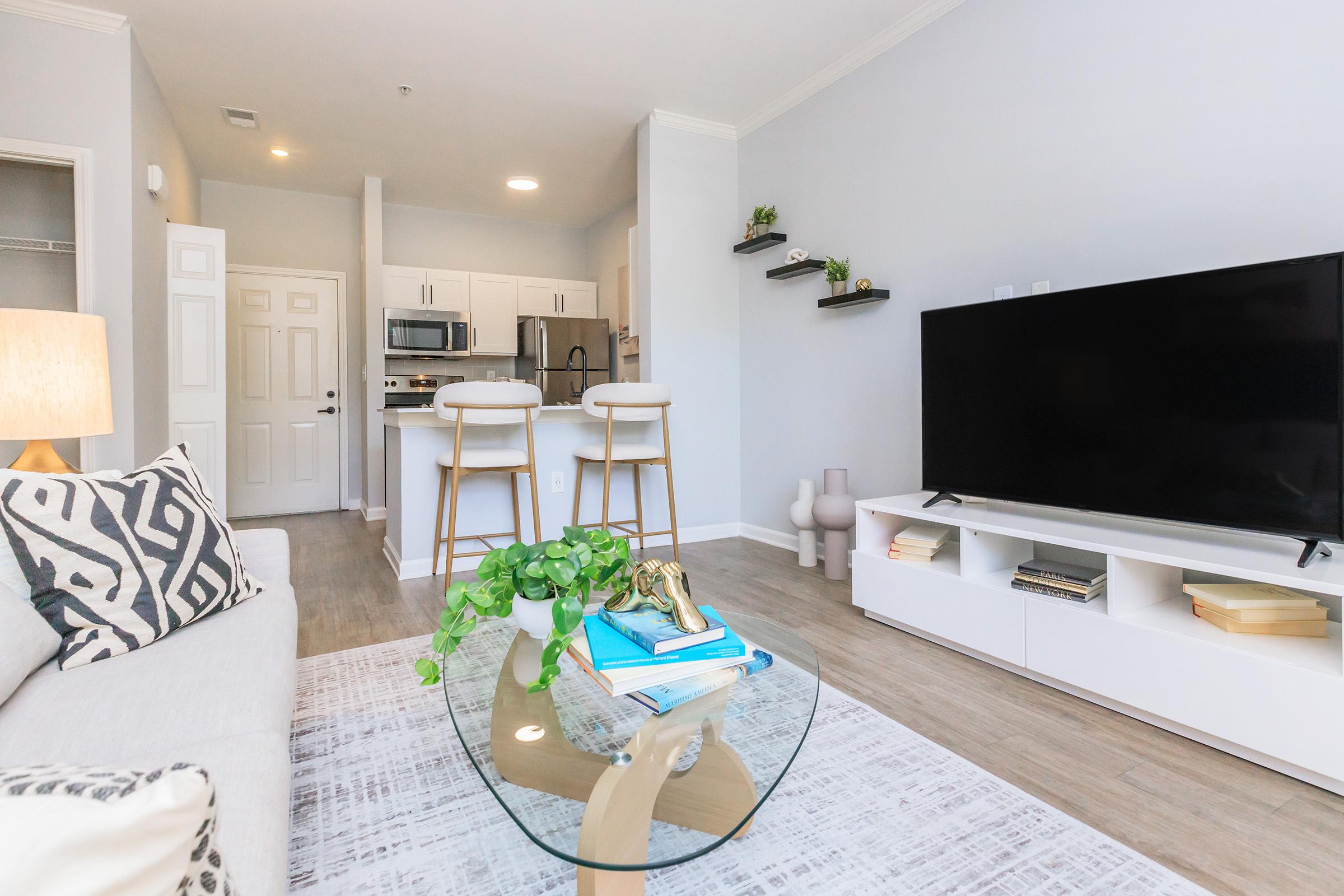Modern living room featuring a light-colored sofa with patterned cushions, a glass coffee table with decorative items, and a large flat-screen TV on a white media console. In the background, there is an open kitchen area with bar stools and shelves adorned with plants. Bright and airy ambiance with neutral tones.