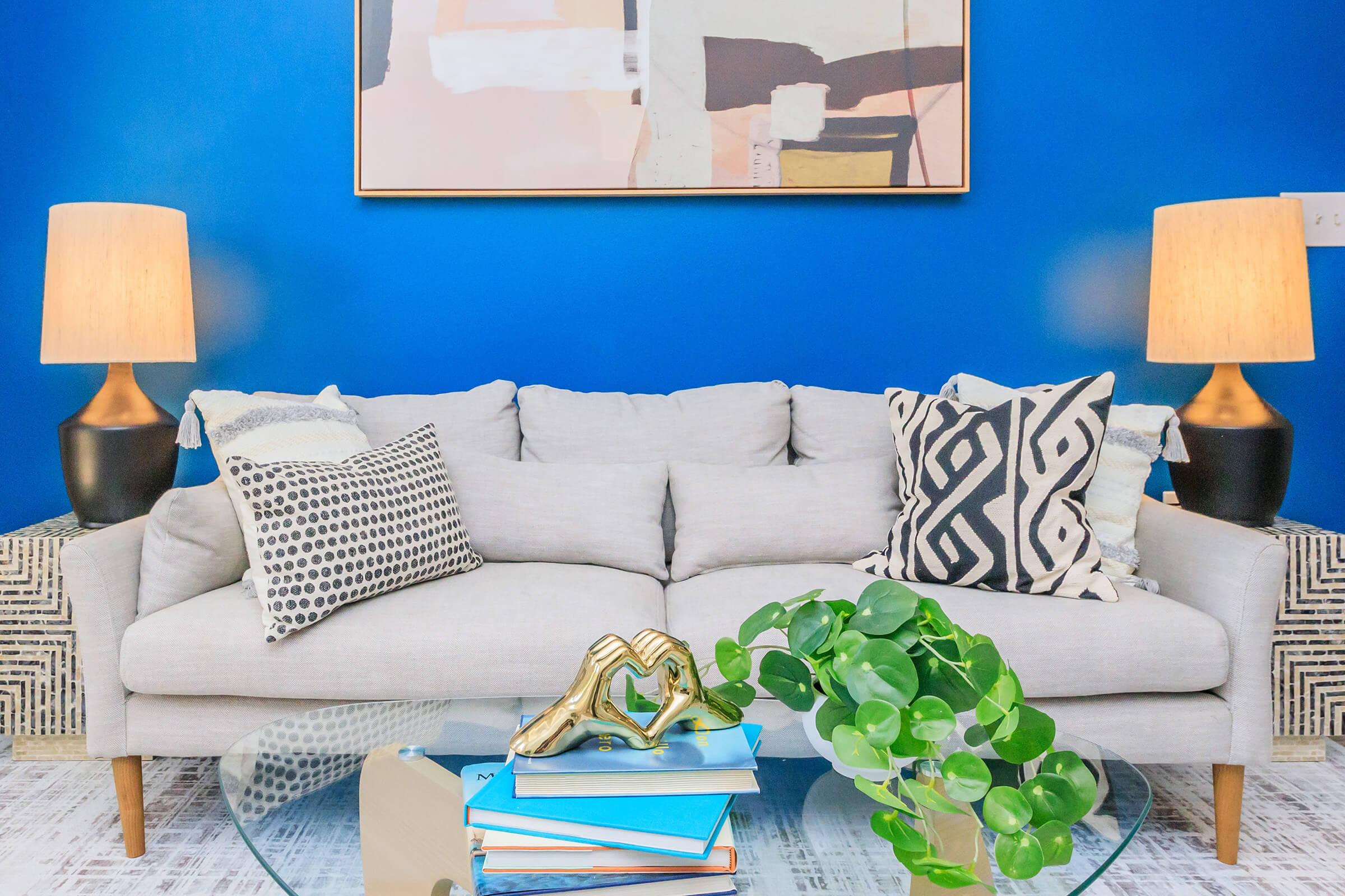 A stylish living room featuring a light gray sofa with decorative pillows, a glass coffee table with stacked books and a small potted plant. The background is a bold blue wall with modern artwork framed above the sofa, and two stylish lamps with patterned bases illuminate the space.
