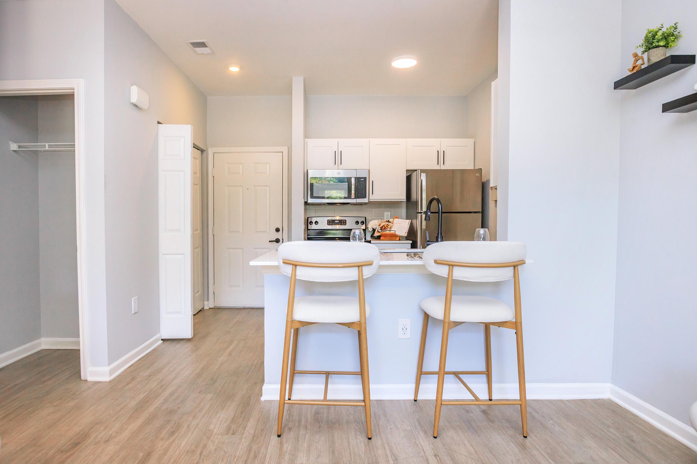 A modern kitchen with white cabinetry and stainless steel appliances. A bar area with two stylish white stools faces the kitchen. The walls are painted in a light gray, and there is a door leading to another room. Natural light fills the space, creating a bright and inviting atmosphere.