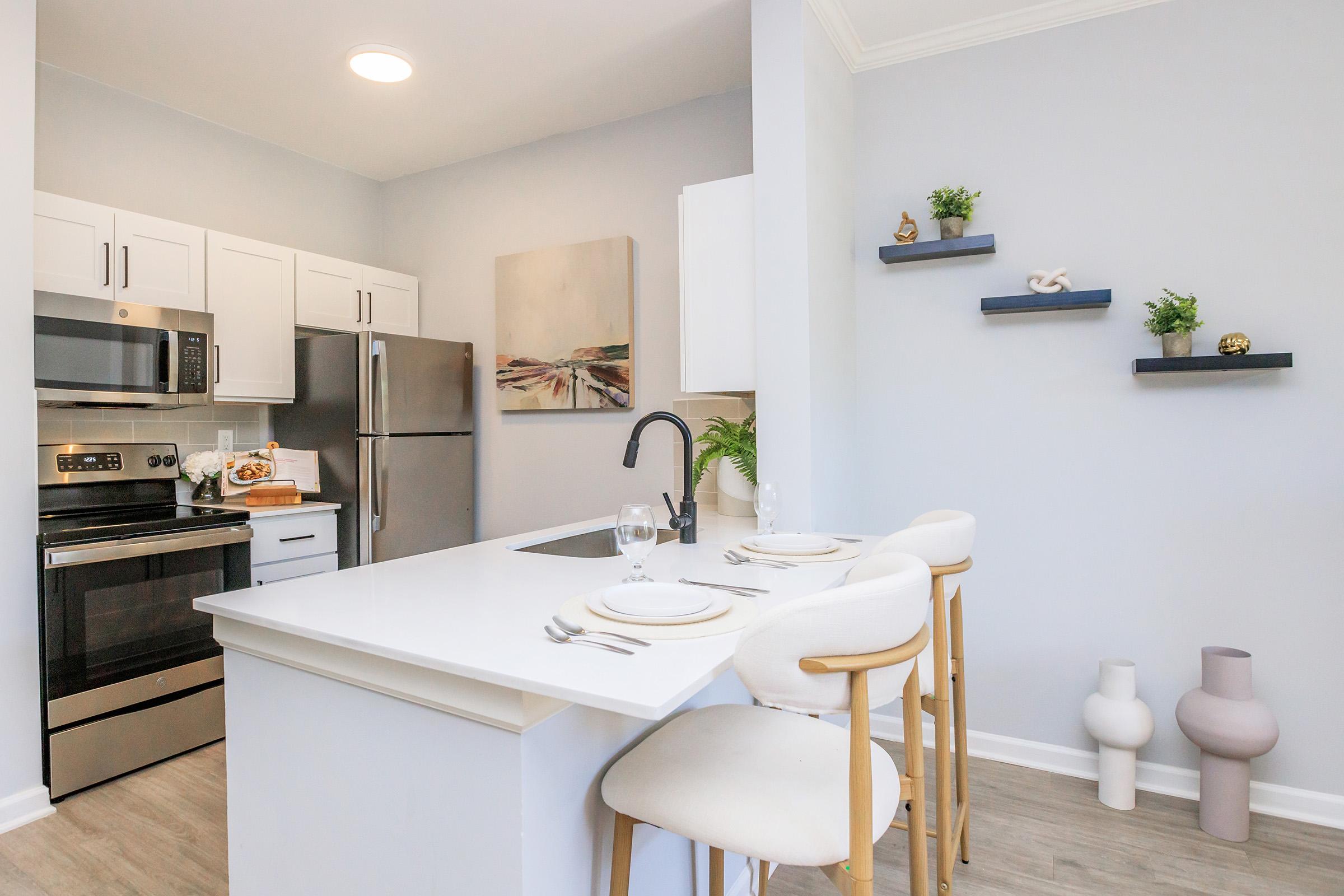 Modern kitchen with white cabinetry, stainless steel appliances, and a center island featuring three white bar stools. Decor includes a small plant and minimalist wall shelves displaying decorative items. Soft gray walls enhance the bright atmosphere. Natural light floods the space through a ceiling fixture.
