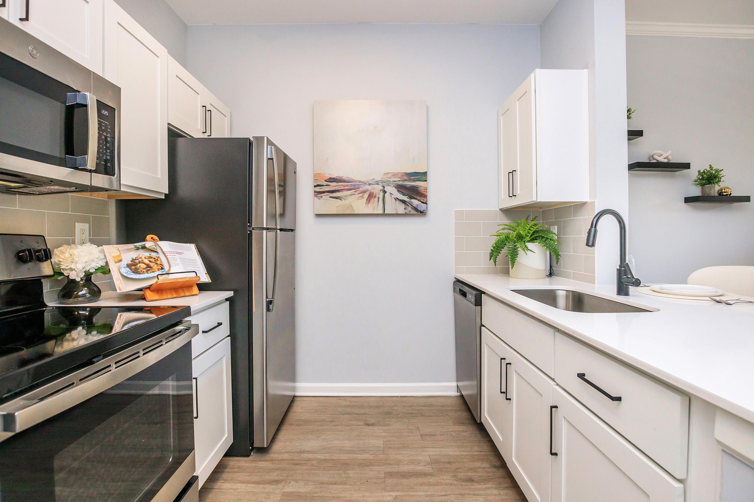 A modern kitchen featuring stainless steel appliances, including a fridge and stove. White cabinets and a sleek countertop provide a clean aesthetic. A colorful abstract painting hangs on the wall, and greenery is placed near the sink. The overall design is bright and inviting.