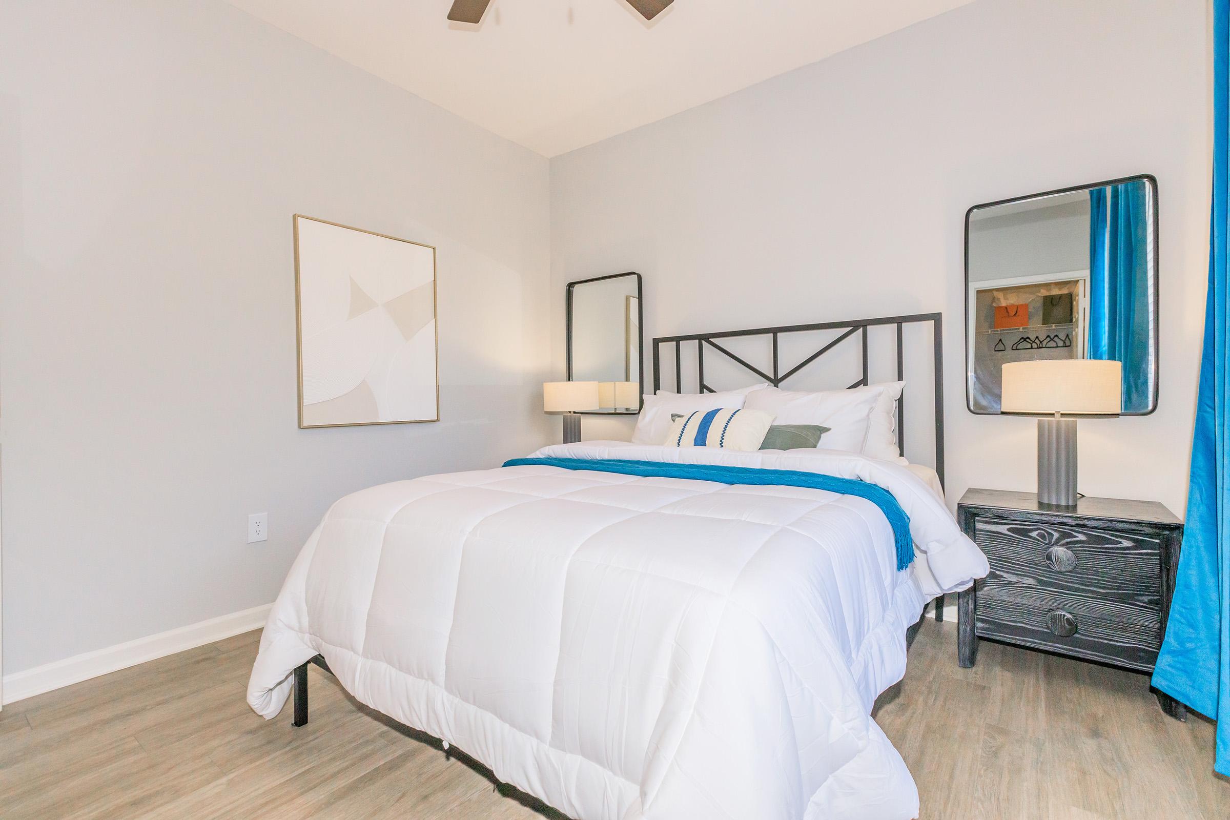 A cozy bedroom featuring a bed with white bedding and blue accents, flanked by two mirrors on nightstands. The room has light gray walls, a ceiling fan, and a piece of abstract art above the bed, creating a modern and inviting atmosphere. Natural light comes in from a window with blue curtains.