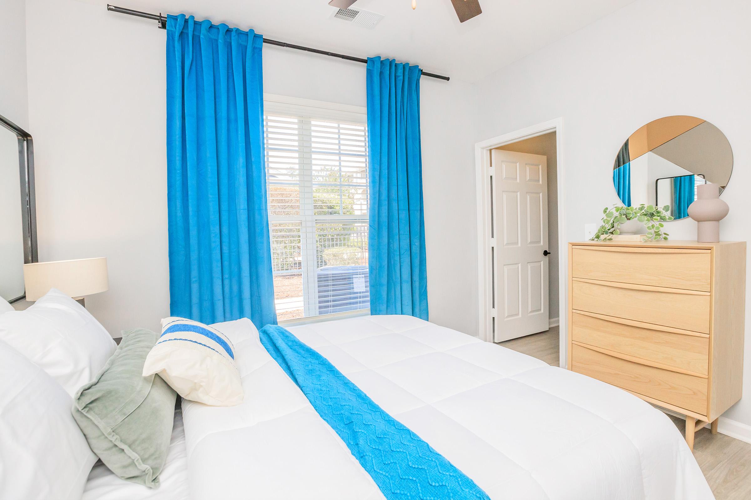 A bright and modern bedroom featuring a white bed with a blue throw blanket, light green pillow, and white linens. Blue curtains frame a window that lets in natural light. A wooden dresser sits beside the bed, and a round mirror is mounted on the wall, enhancing the room's airy and contemporary feel.