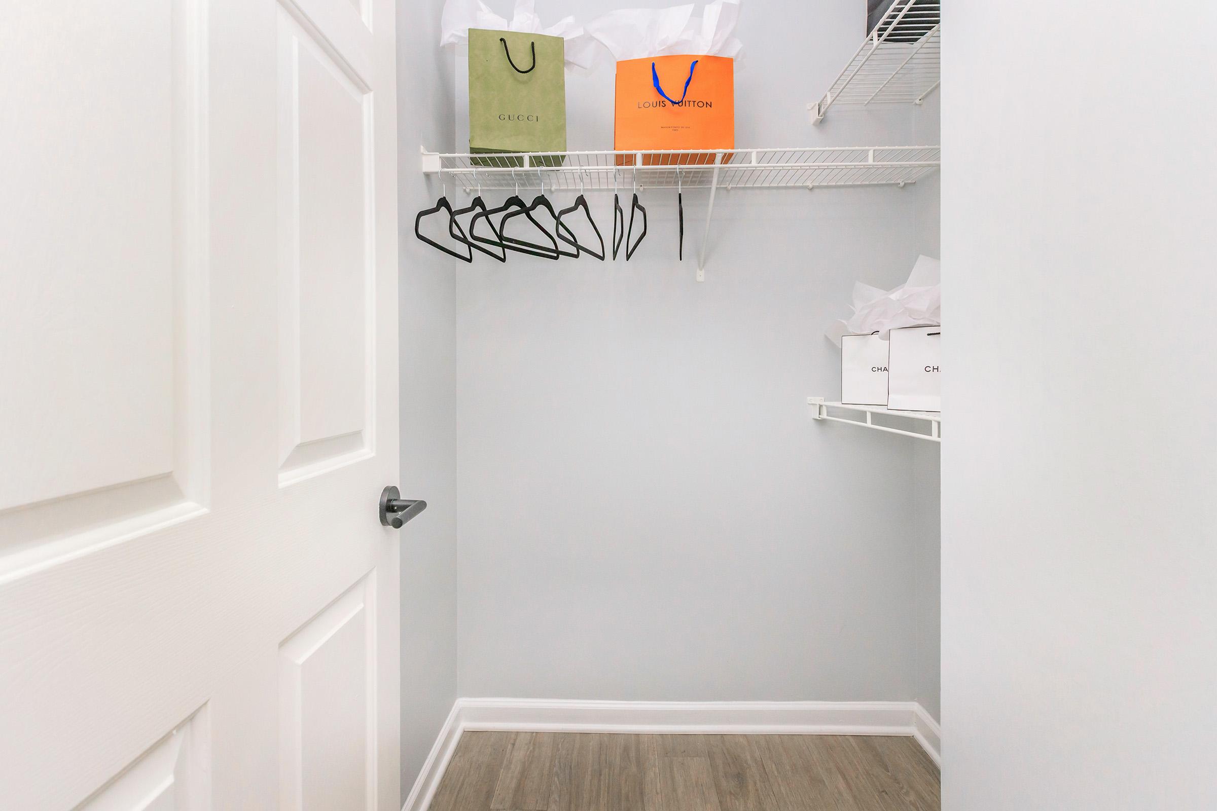 A well-organized closet featuring a white door, gray walls, and a wooden floor. On the left, several black hangers are hanging from a wire shelf. On the shelf are shopping bags from luxury brands, including green and orange bags, along with white bags, suggesting a stylish and minimalistic storage space.