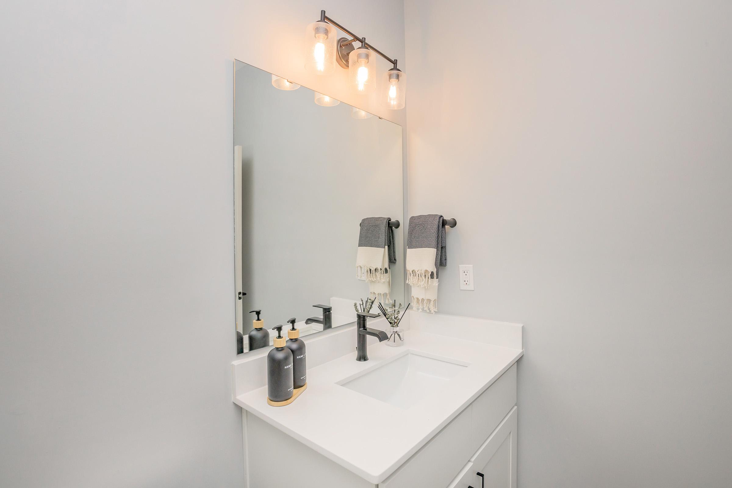 A modern bathroom vanity with a white countertop and sink. Above the sink, there is a large mirror flanked by three light bulbs. A gray hand towel is hanging on a stylish towel holder. On the countertop, there are two bottles, one dark and one light, and a decorative plant in a small vase.