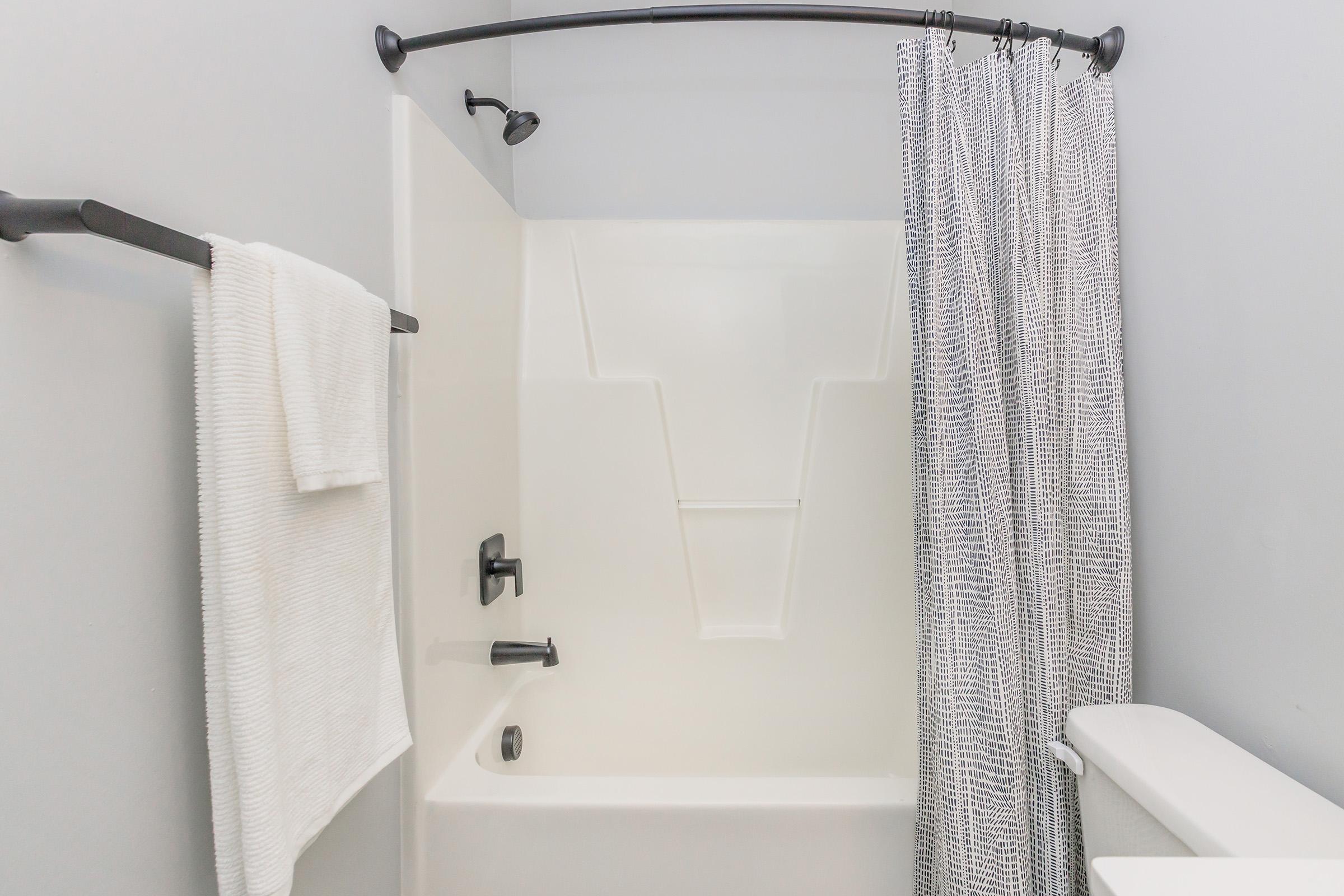 A modern bathroom featuring a white bathtub with a showerhead, a gray patterned shower curtain, and a towel hanging on a black towel bar. The walls are painted light gray, creating a clean and minimalist aesthetic. The space is well-lit and organized.