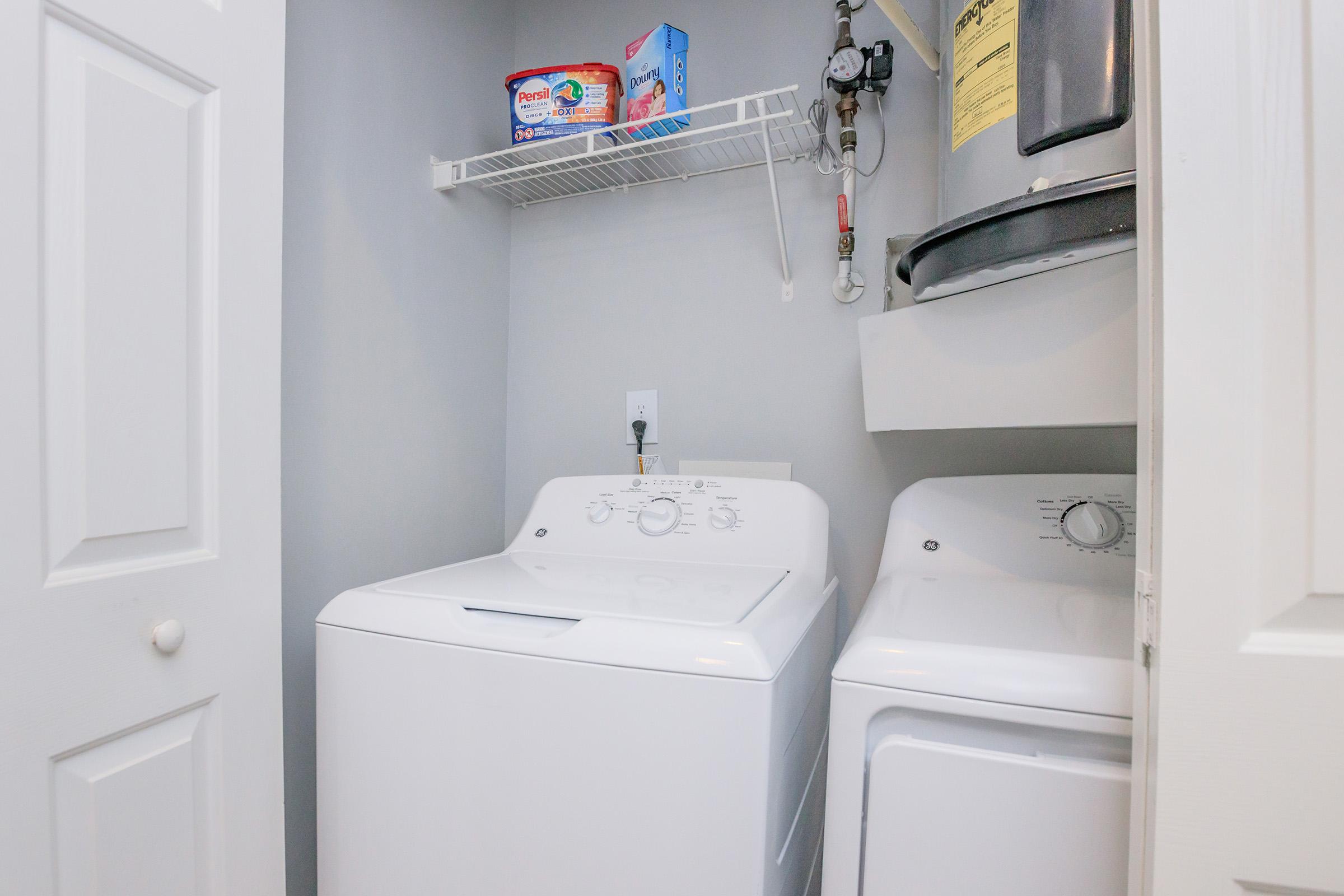 A laundry room features a white washing machine and a dryer side by side. There is a shelf above with laundry detergent and fabric softener. The room has light gray walls and a water heater is visible beside the appliances. The door is partially open, revealing the organized space.