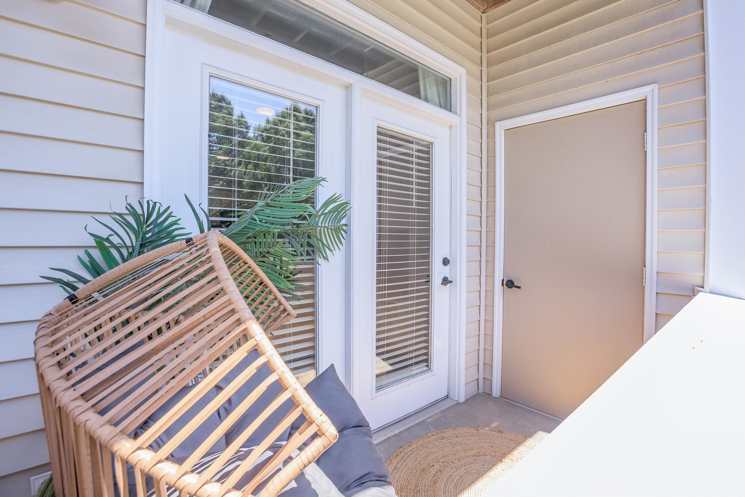 An inviting entryway with two glass-paneled double doors featuring horizontal blinds. To the side, a solid beige door leads to another room. A wicker chair is tipped over against a wall, and a potted green plant adds a touch of nature to the space. The exterior has light-colored siding and a small rug on the floor.