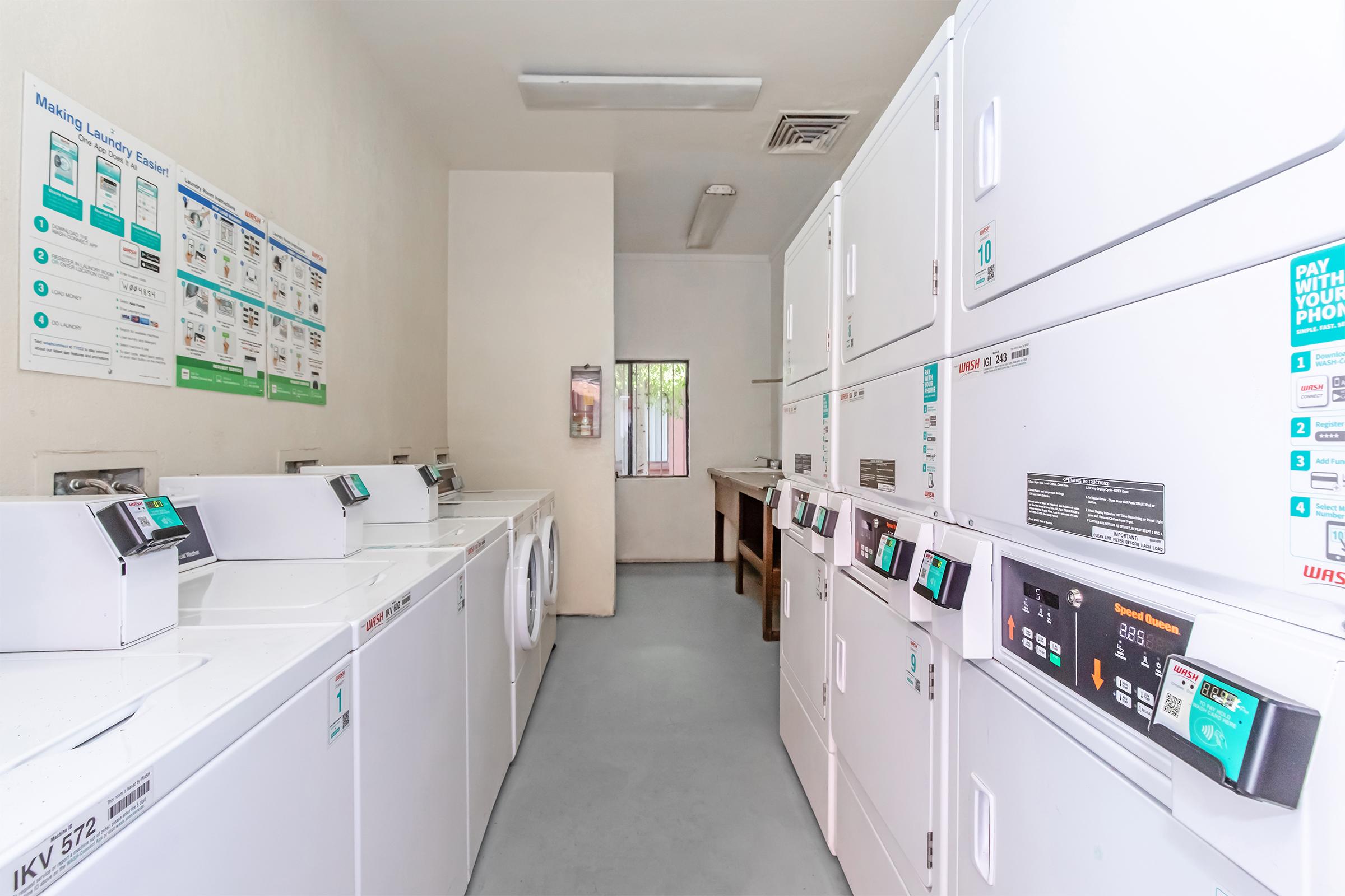 A laundry room featuring multiple white washing machines and dryers, arranged in a row. The walls display instructional signs and information about laundry services. A large window at the far end allows natural light, and a folding table is visible on the right side of the room.