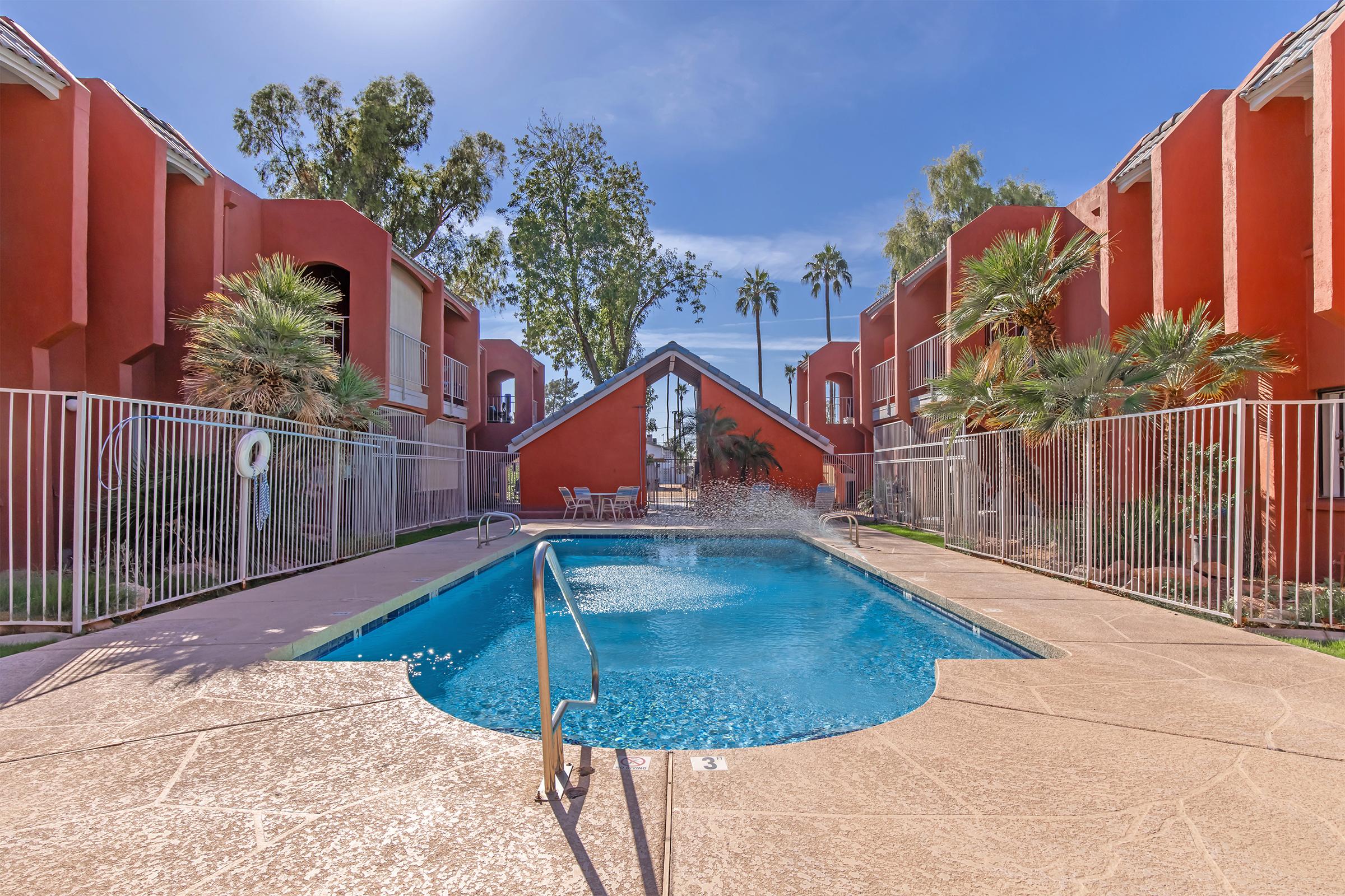 A bright, sunny pool area surrounded by red apartment buildings. The clear blue water of the pool features a splash from a fountain. Palm trees and greenery add a tropical feel, while lounge chairs are positioned nearby. A metal fence encloses the pool for safety.