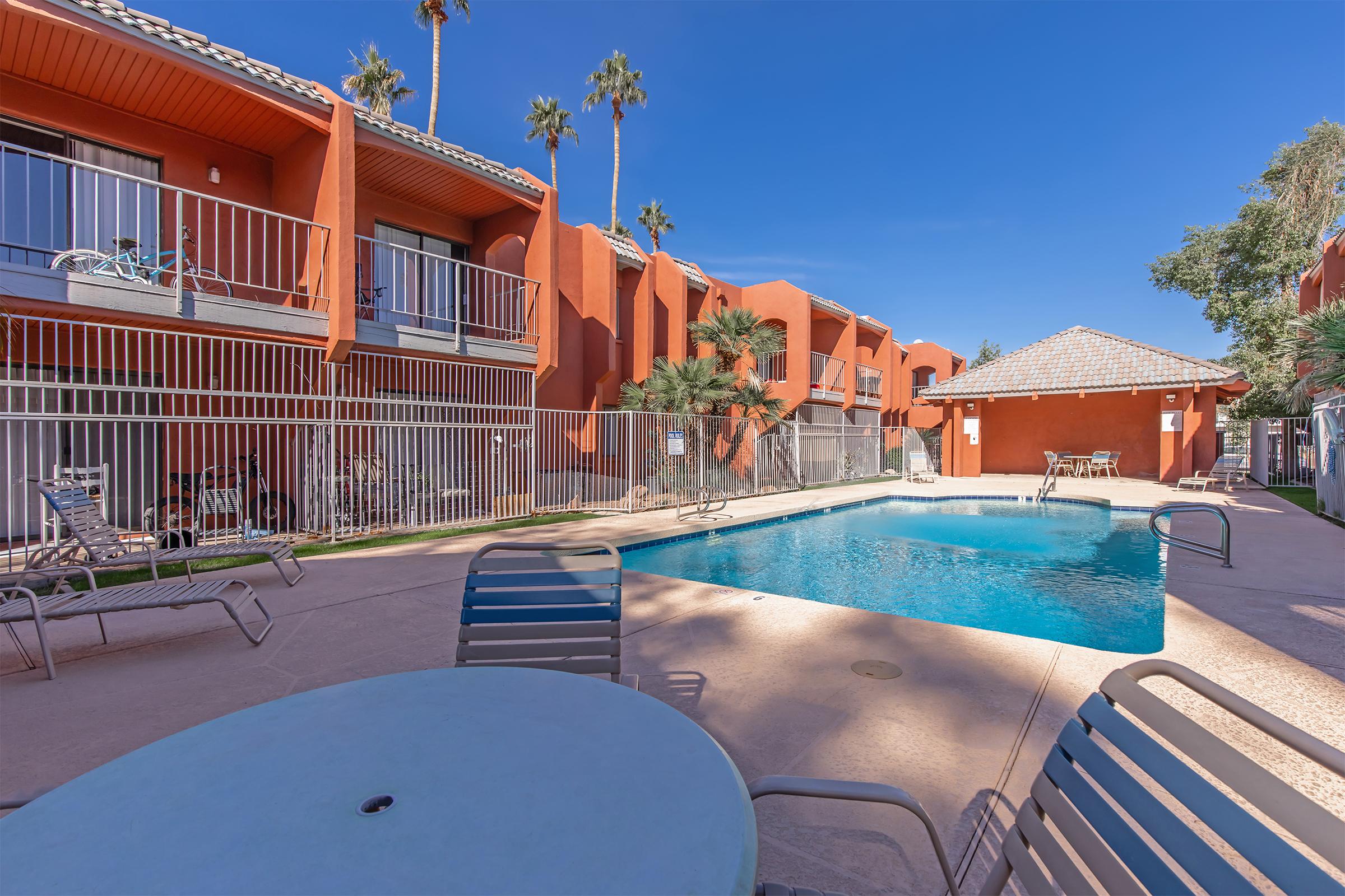 A bright, sunny view of a swimming pool surrounded by lounge chairs and tables, set against a backdrop of orange-colored apartment buildings and palm trees. The clear blue sky enhances the inviting atmosphere of the outdoor space.