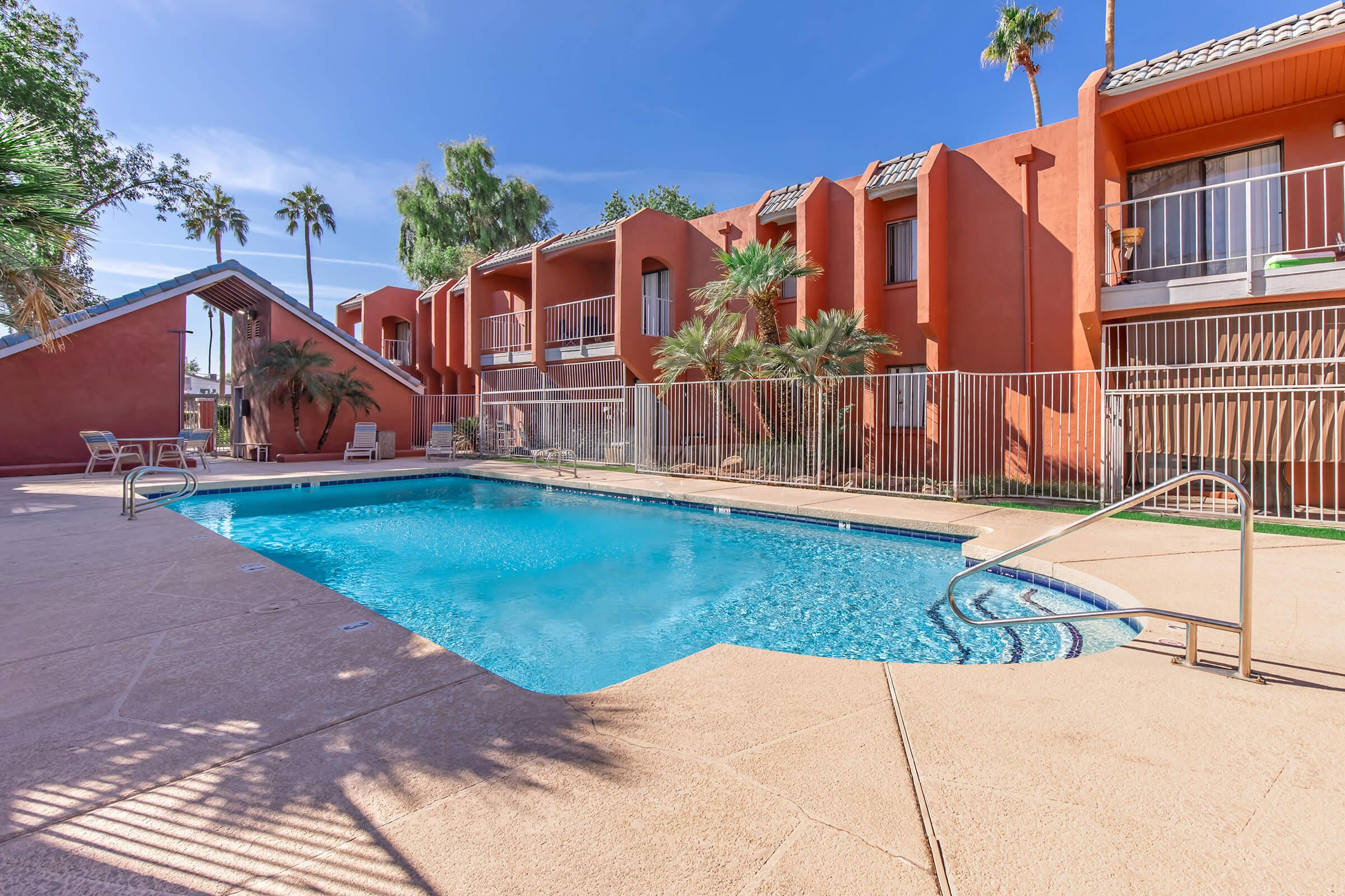 A sparkling blue swimming pool surrounded by a paved deck, with lounge chairs nearby. Colorful palm trees and the orange exterior of a resort-style building can be seen in the background under a clear blue sky, creating a relaxing outdoor atmosphere.