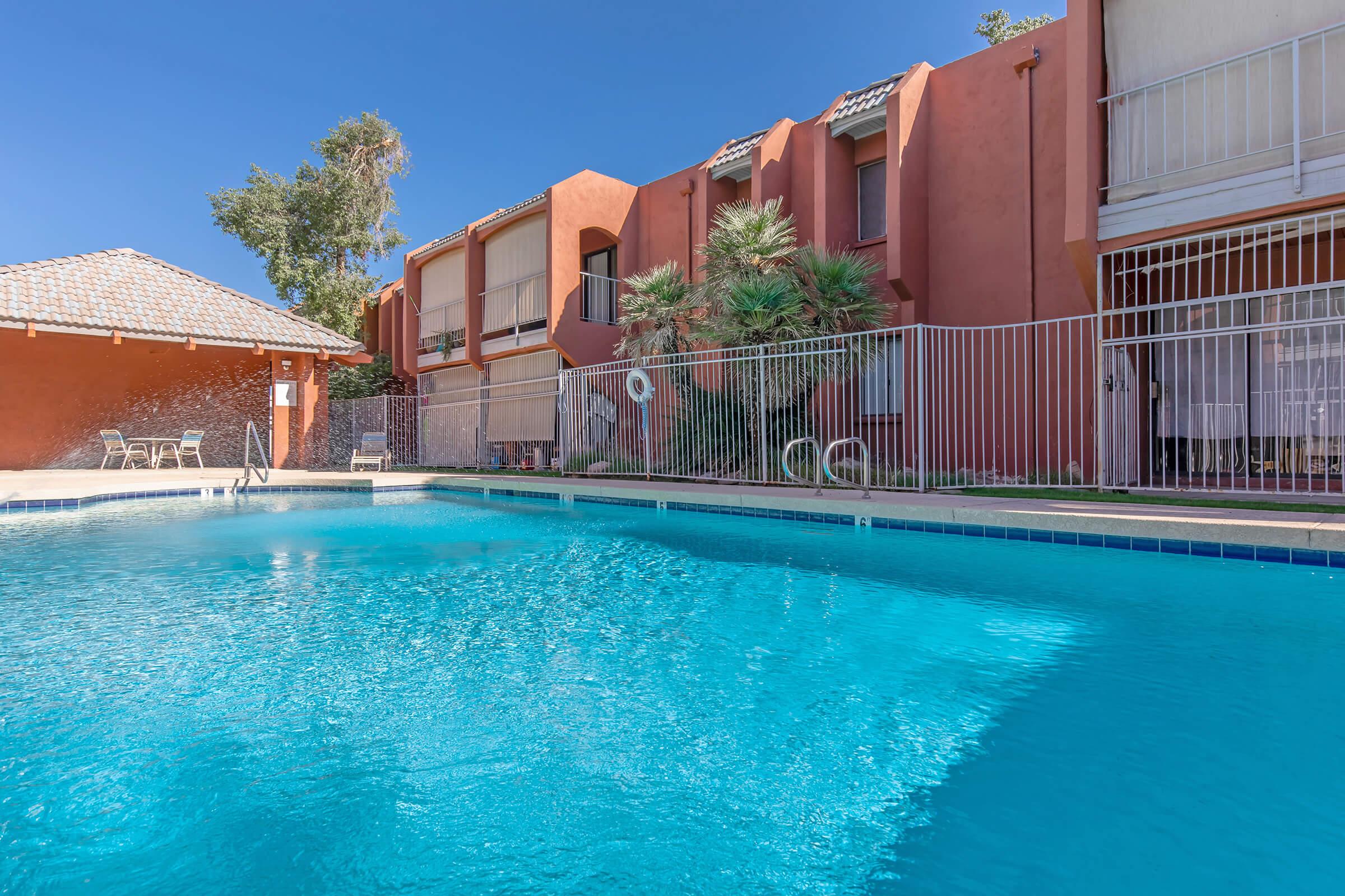 A clear blue swimming pool surrounded by a fence, with an orange-hued building in the background. The area features palm trees and lounge chairs, under a sunny blue sky. The pool area is well-maintained and inviting.