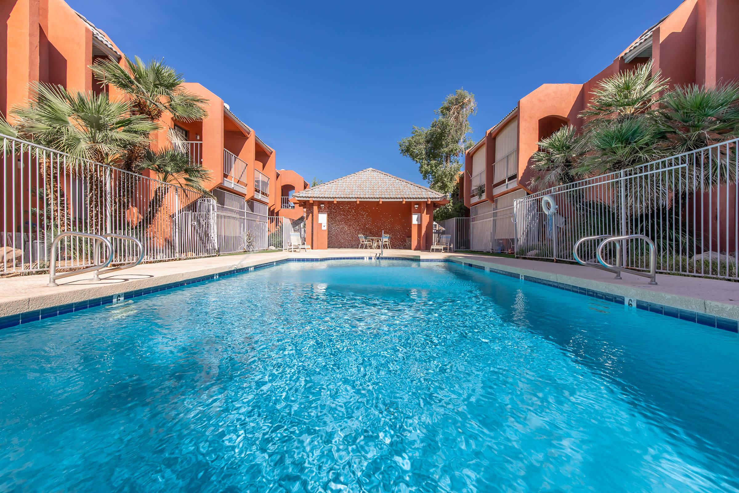 A clear swimming pool surrounded by lush palm trees and two orange buildings with balconies on either side. In the background, there's a small house or facility, and the sky is bright blue, creating a serene and inviting atmosphere.