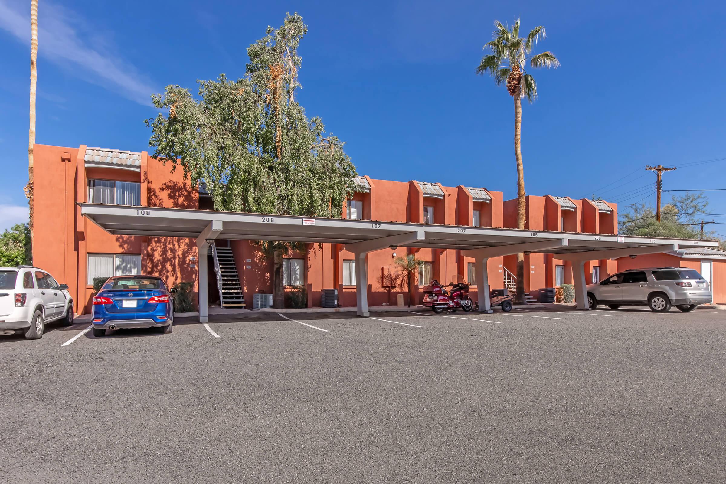 A two-story, orange-colored building with multiple covered parking spaces in front. There are several vehicles parked, including a blue car and motorcycles. Palm trees are visible, and the sky is clear and blue, indicating a sunny day. The parking lot has clearly marked spaces.