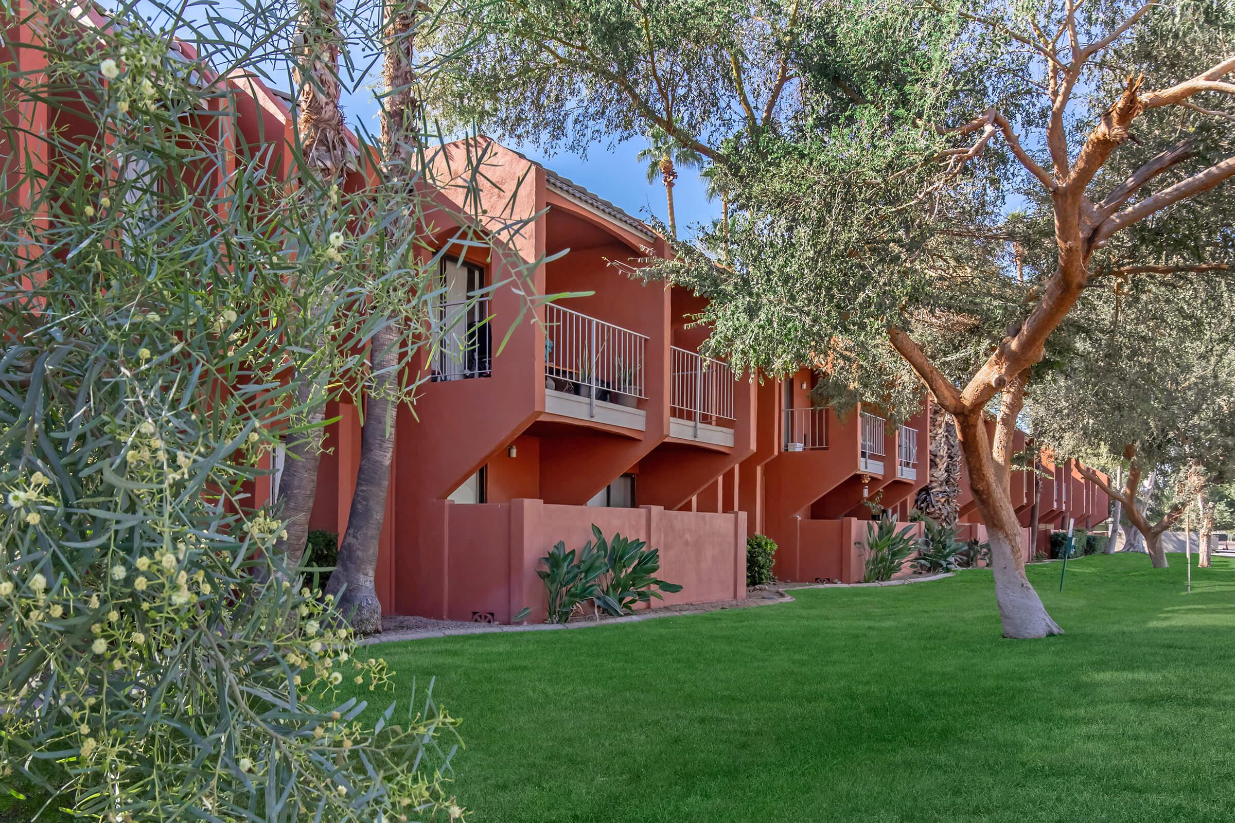 A row of modern, terracotta-colored apartments surrounded by lush green grass and trees. The buildings feature balconies and are set against a clear blue sky, creating a vibrant and inviting atmosphere. The surrounding foliage adds to the serene ambiance of the area.