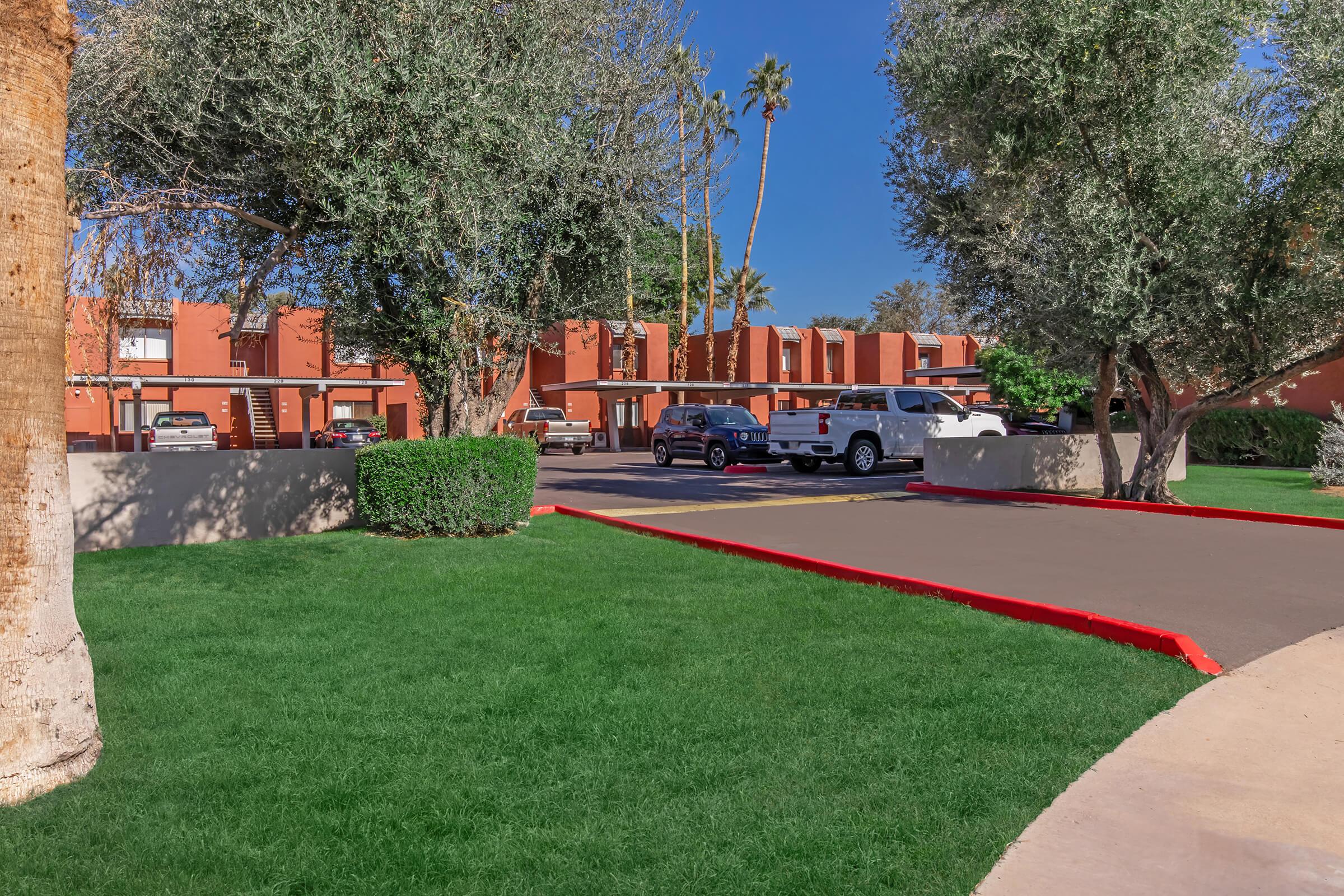 A view of a well-maintained parking area surrounded by lush green grass and palm trees. The scene features brown buildings in the background and several parked vehicles, including a white truck. Clear blue skies and a sunny atmosphere indicate a warm climate.