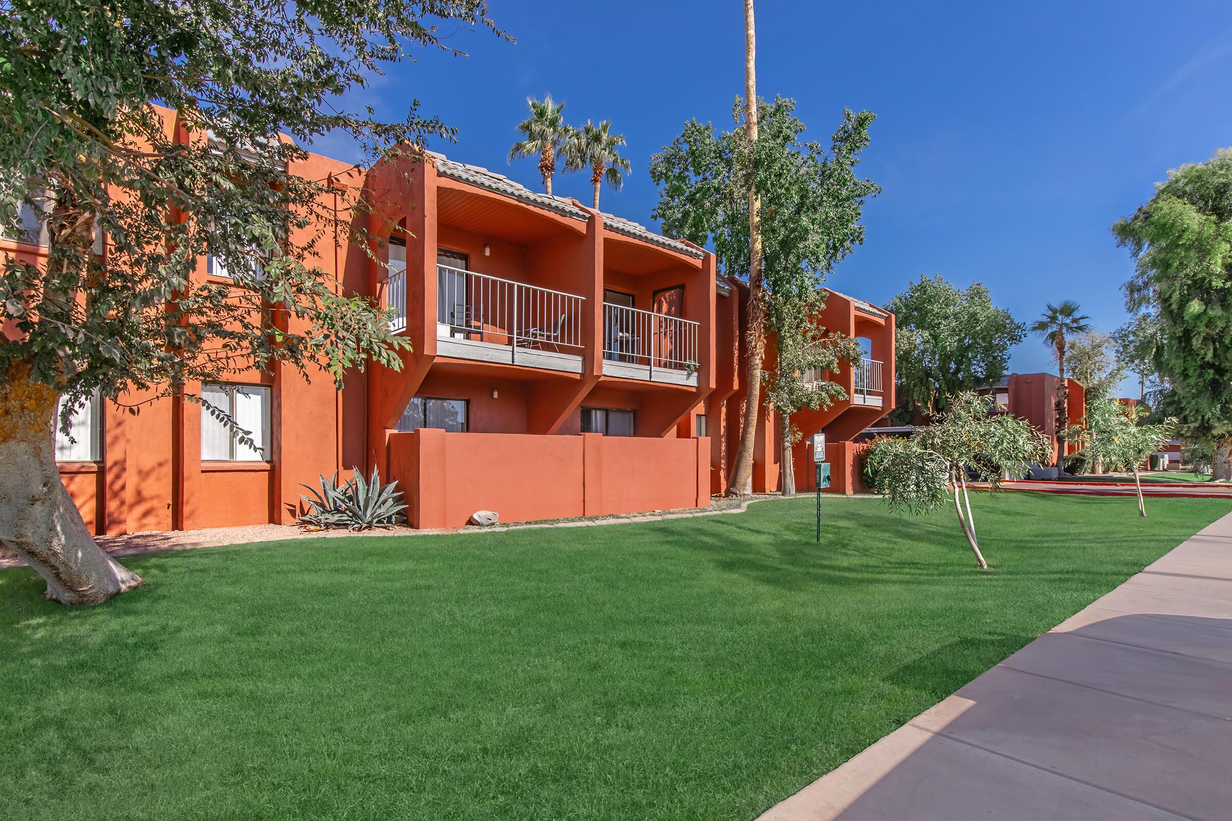 Colorful orange buildings with balconies surrounded by green grass and palm trees under a clear blue sky. Pathway in the foreground leads through the landscaped area, creating a peaceful outdoor setting.