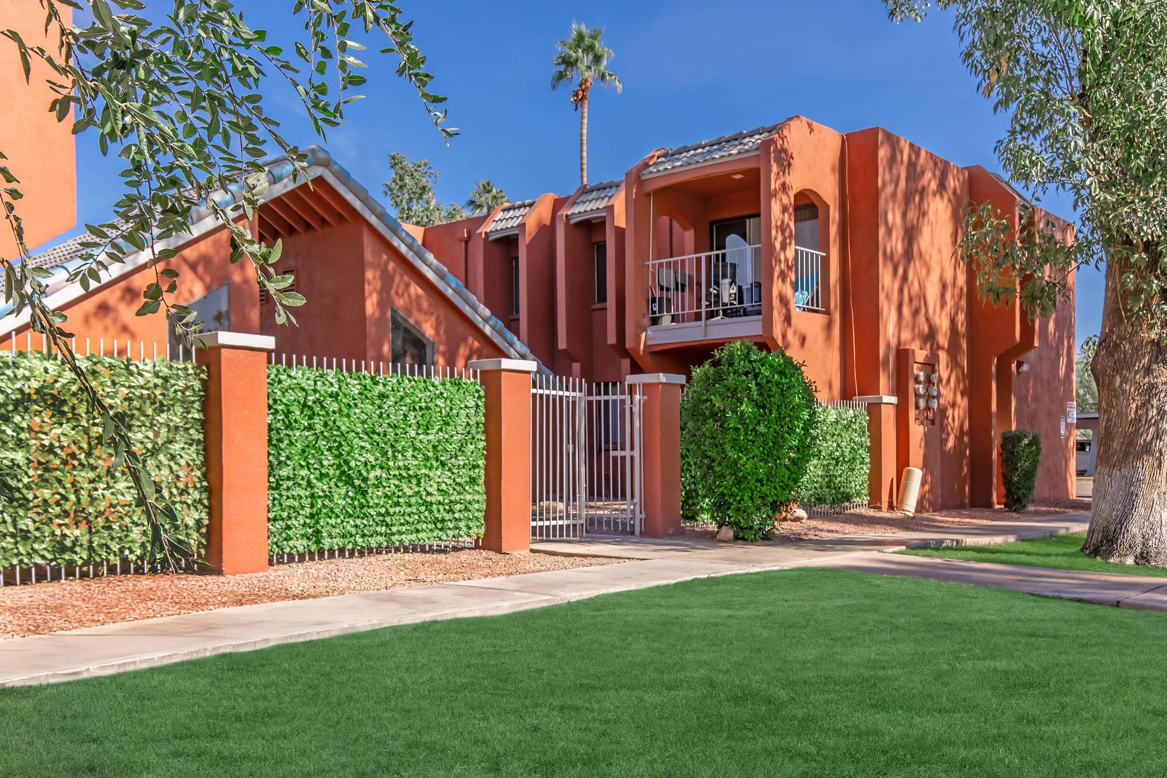 A vibrant orange apartment building surrounded by lush greenery and a manicured lawn. The structure features balconies and is enclosed by a decorative metal gate. Tall palm trees are visible against a clear blue sky, contributing to a sunny, inviting atmosphere.