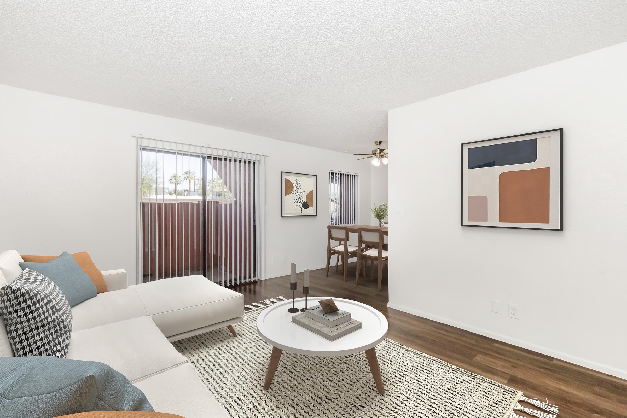 A bright and modern living room featuring a light-colored sectional sofa with decorative pillows, a round coffee table with books and decor items, and a textured area rug. Large windows with blinds allow natural light, and a dining area with a wooden table and chairs is visible in the background. Wall art adds a contemporary touch.