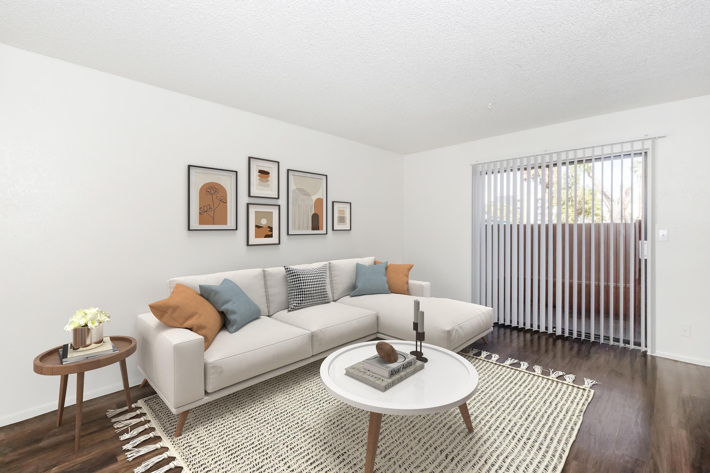 A modern living room featuring a light-colored sectional sofa with decorative pillows, a round coffee table with a decorative tray, and a small side table with a vase of flowers. The walls are adorned with framed artwork, and there is a set of sliding glass doors with vertical blinds leading to an outdoor area.