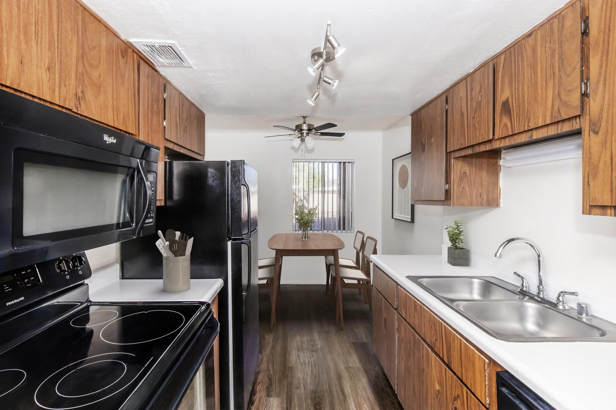 A modern kitchen featuring wooden cabinets, a black refrigerator and stove, and a double sink. There is a dining table with chairs, natural light coming through a window, and decorative plants on the countertop. The overall design is clean and functional.