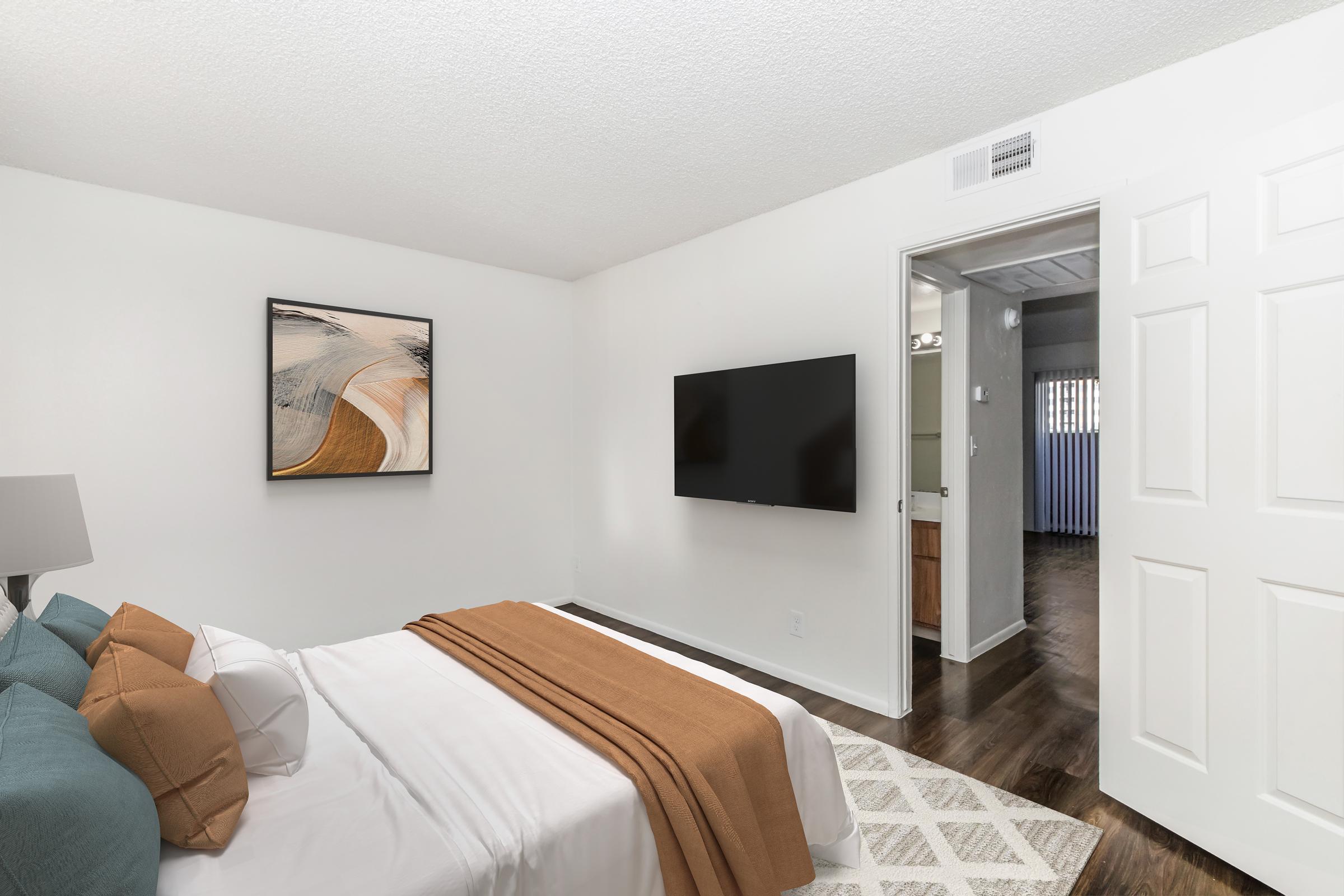 A cozy bedroom featuring a neatly made bed with white linens and an orange throw blanket. There's a modern abstract artwork on the wall, a large flat-screen TV mounted opposite the bed, and an open door leading to a bathroom area. The floor is dark wood, and the room has a bright, inviting atmosphere.