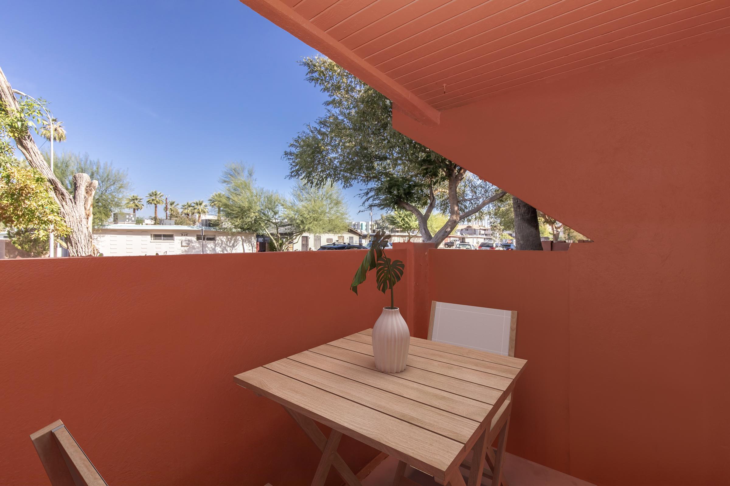 A cozy outdoor patio featuring a light wood table and white chairs against a vibrant orange wall. A simple white vase with a green plant sits on the table. In the background, palm trees and blue skies are visible, creating a serene and inviting atmosphere.