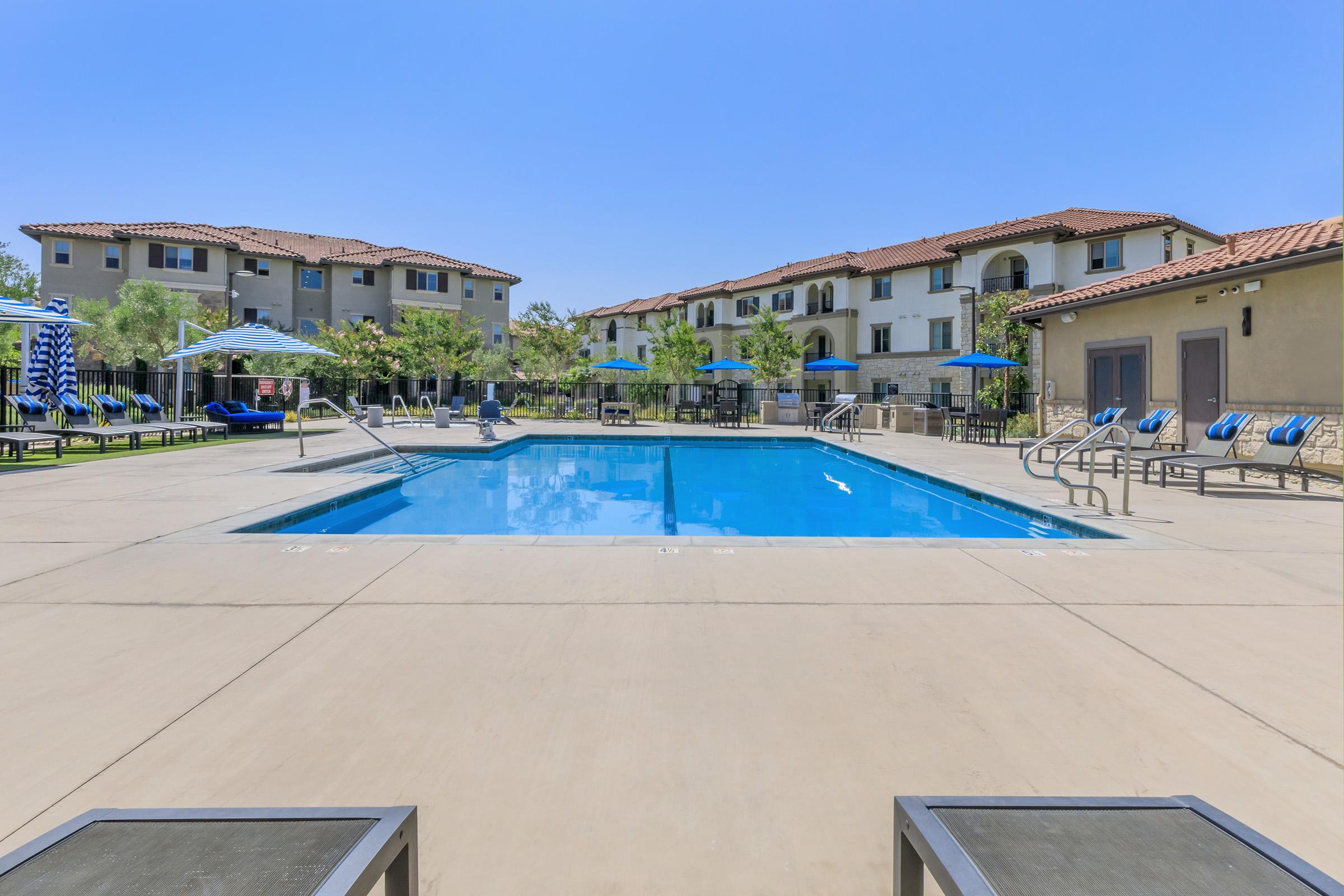 A clear swimming pool surrounded by lounge chairs and umbrellas, set in a residential complex. In the background, multiple multi-story buildings with balconies and landscaping. The sky is blue and clear, indicating a sunny day.