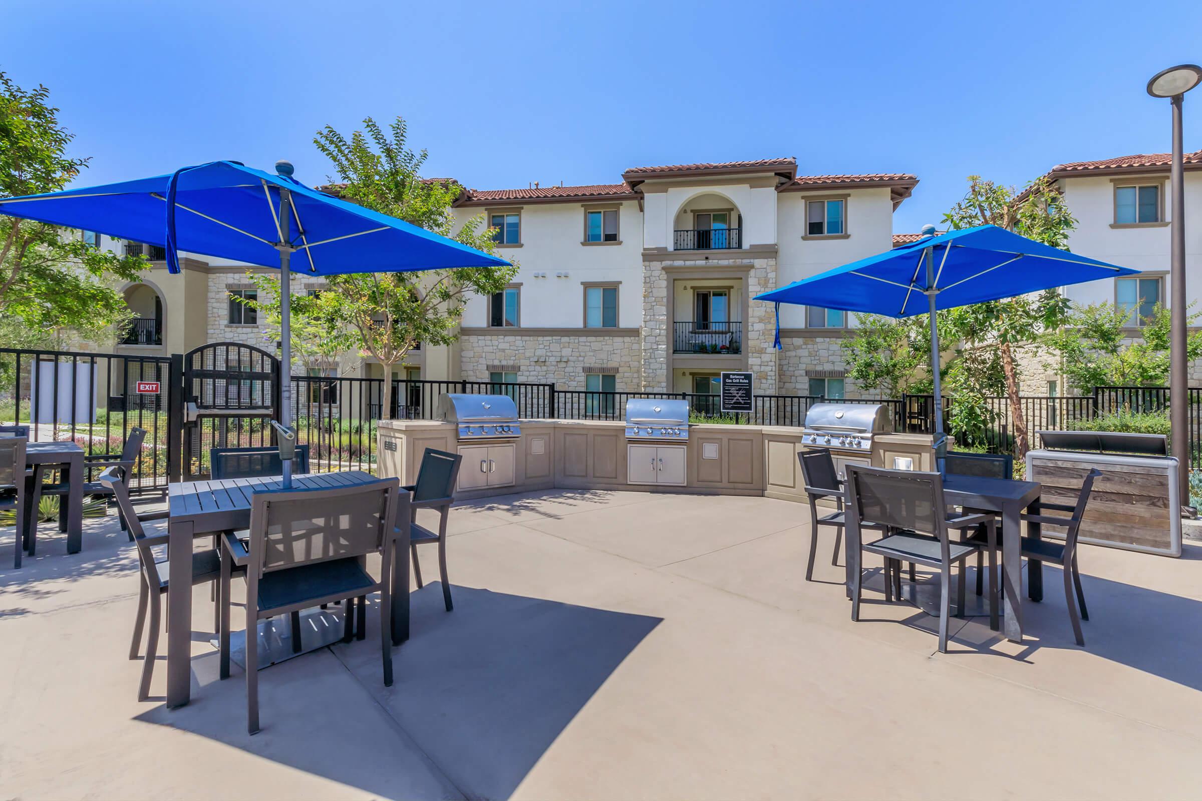 Outdoor area featuring several grill stations and tables under blue umbrellas, with a decorative wall and building in the background. The space is well-lit and surrounded by greenery, creating a welcoming environment for residents to gather and enjoy outdoor cooking and dining.