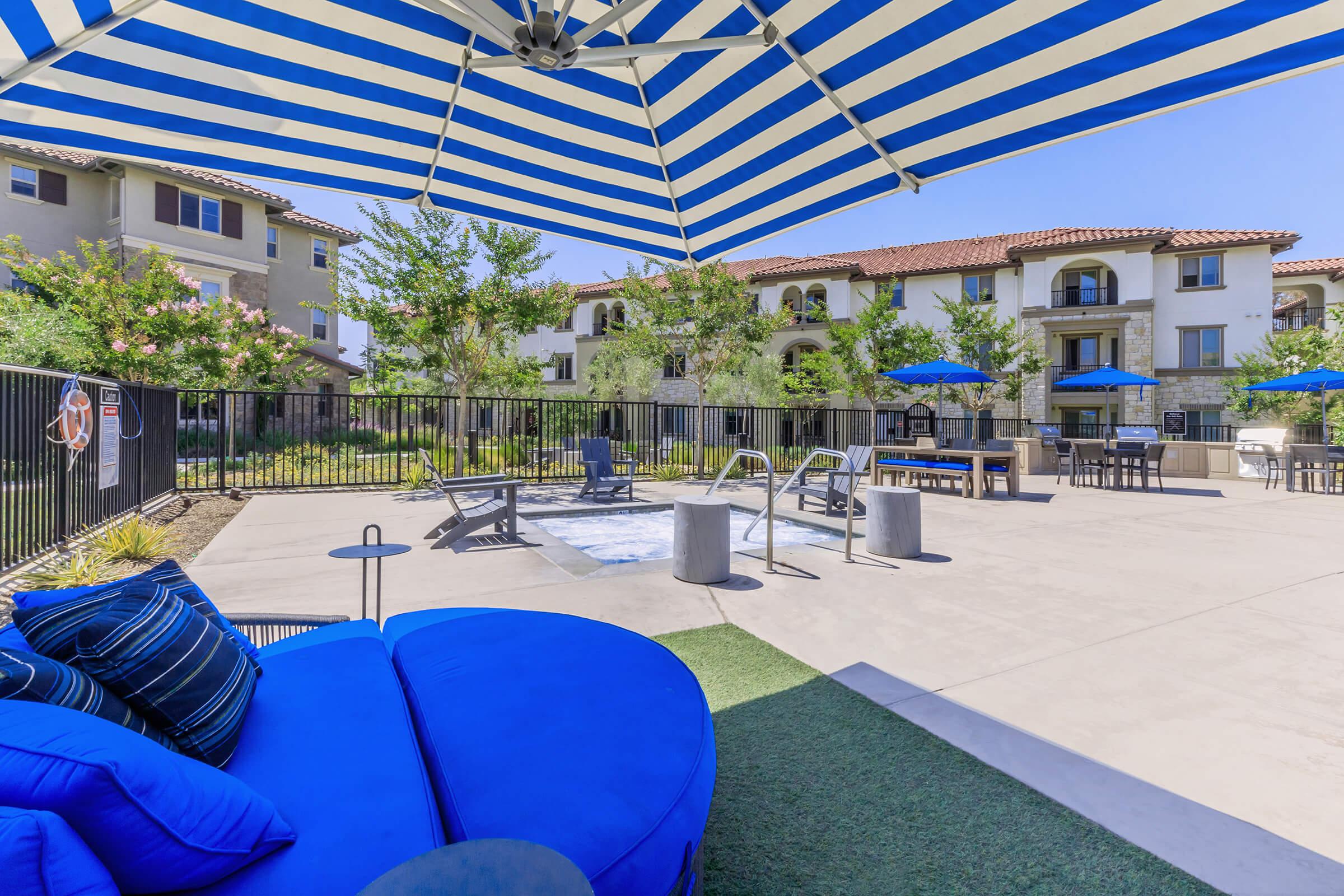 A cozy outdoor area featuring a blue and white striped umbrella, with lounge chairs, a hot tub, and a seating area. Surrounding greenery and amenities enhance the inviting atmosphere, providing a relaxing space near residential buildings. Bright sunny day with clear blue skies.