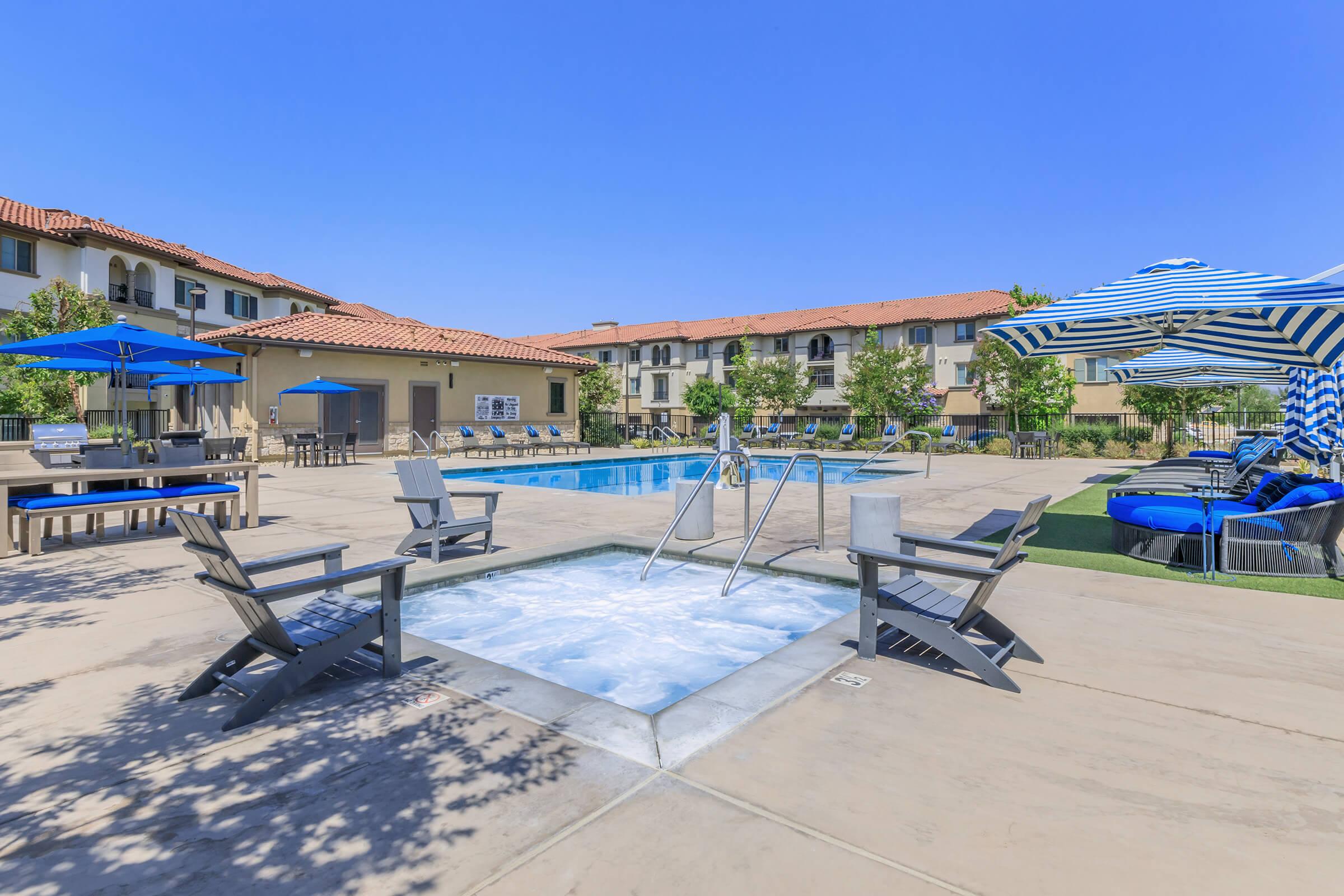 A bright, sunny pool area featuring a large swimming pool surrounded by lounge chairs. There is a hot tub in the foreground, along with patio umbrellas and shaded seating areas. The backdrop includes residential buildings with terracotta roofs and well-maintained landscaping, all under a clear blue sky.